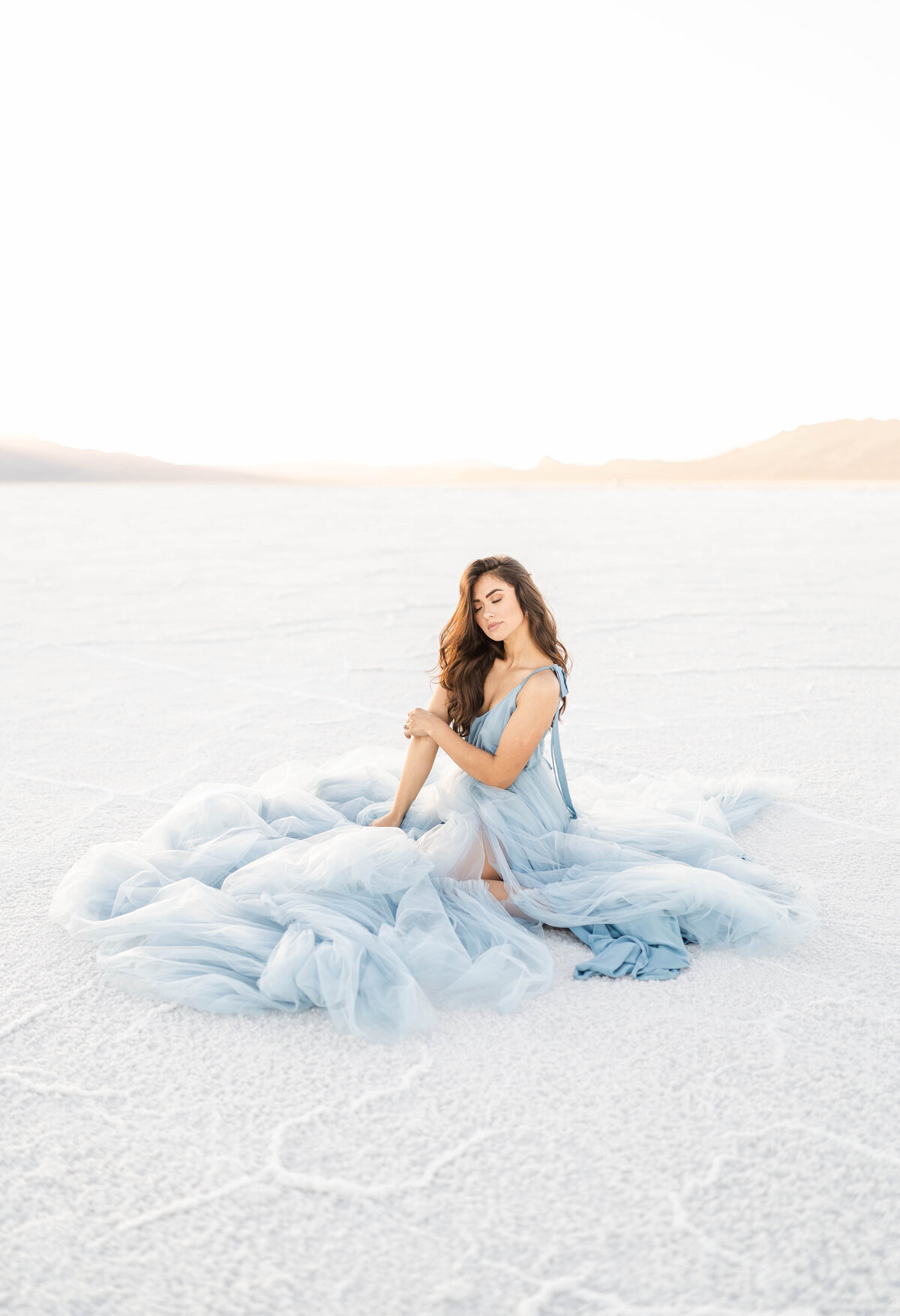 A woman sits in white sand wearing a luxurious soft blue gown glowing in the sunlight photographed by Bay area photographer, Light Livin Photography.
