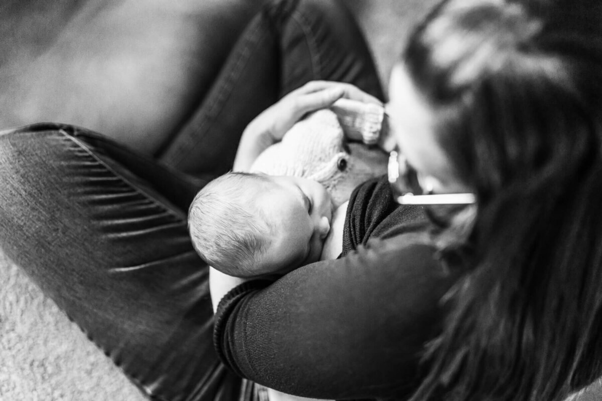 A black and white image of a mother sitting cross legged on her living room floor while her baby breastfeeds.