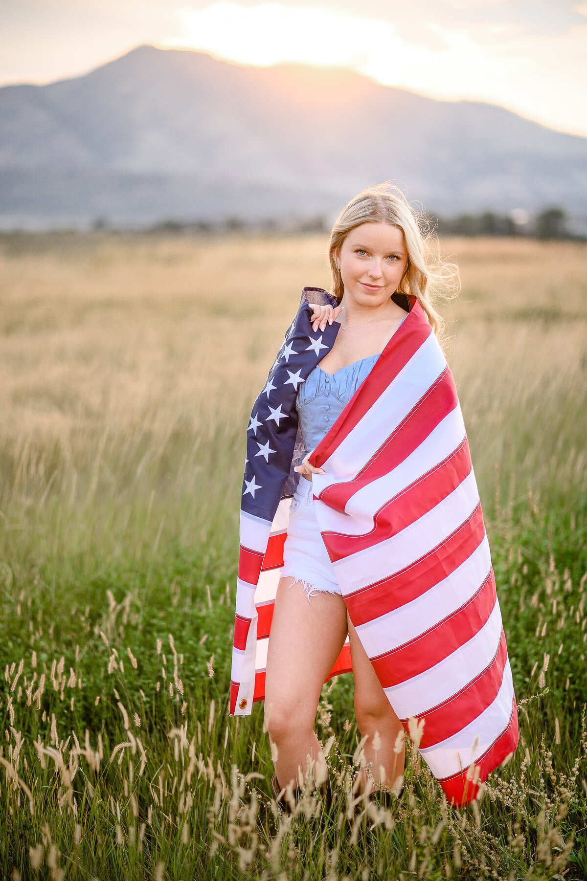 senior pictures with girl wearing a USA flag around her shoulders in a field  with a mountain in the distance captured by denver senior photographer