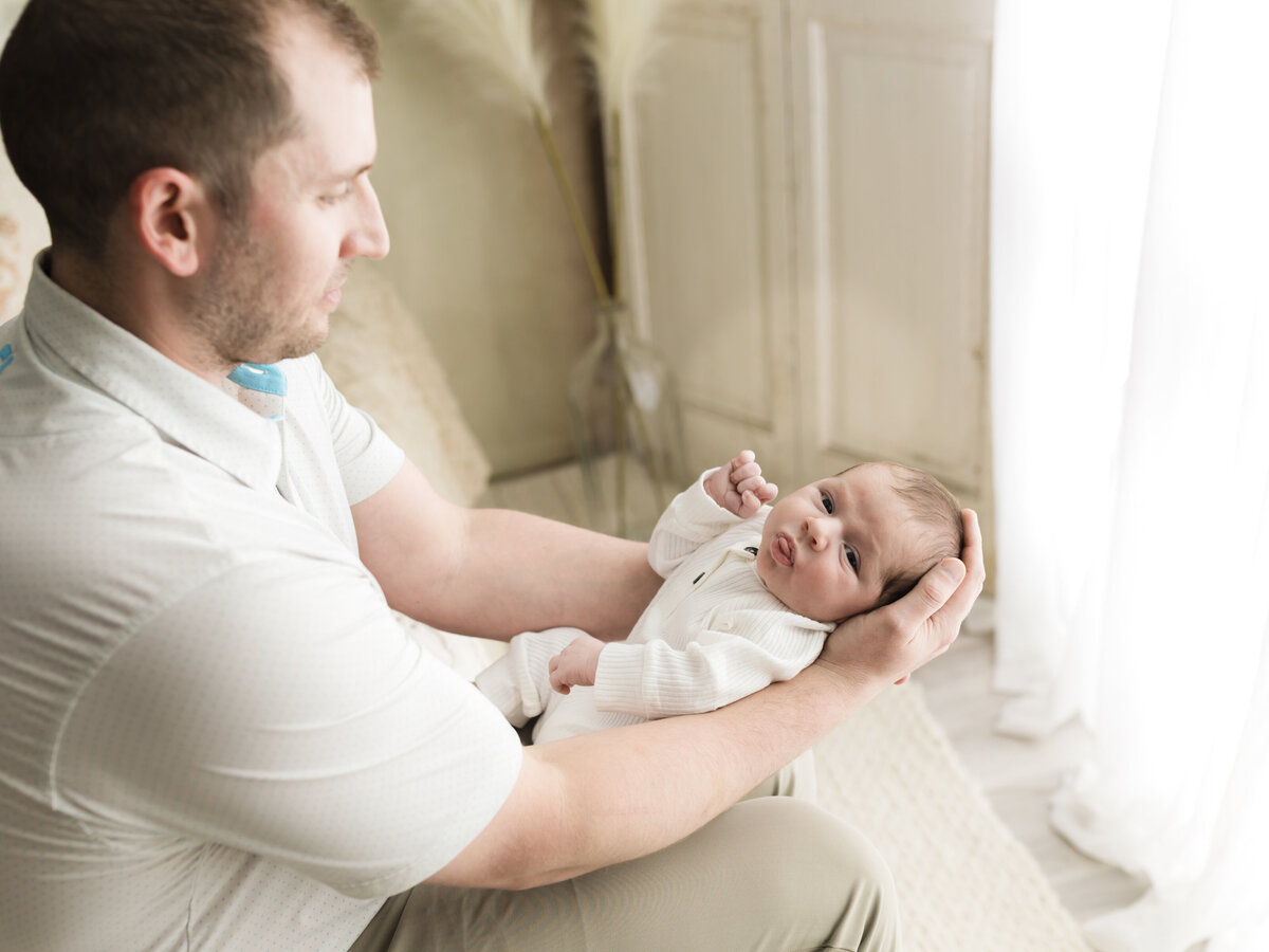 dad holding newborn baby boy in white onesie for studio photoshoot 
