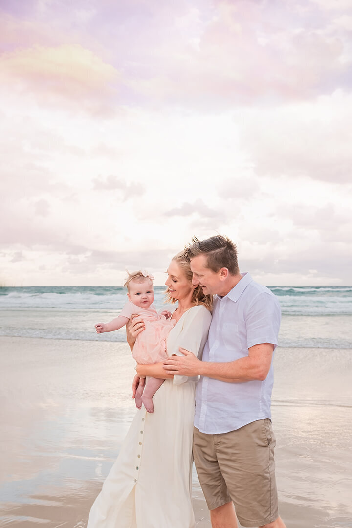 Light and airy family photo on a Brisbane beach: Mum and dad holding baby with joy