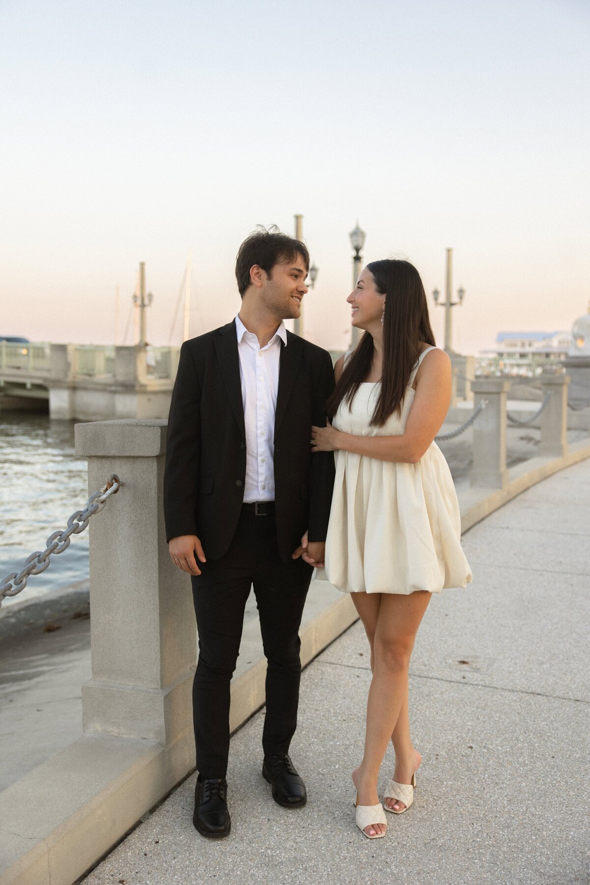 A couple strolls hand-in-hand along the bayfront in St. Augustine, Florida, during their engagement session. The bride-to-be wears a chic white mini dress, while her fiancé is dressed in a black suit with a white shirt. They smile at each other against a backdrop of soft evening light, sailboats, and historic lampposts. Captured by Heartful Films, a Jacksonville engagement photographer and videographer known for stylish, natural portraits in iconic Florida locations.