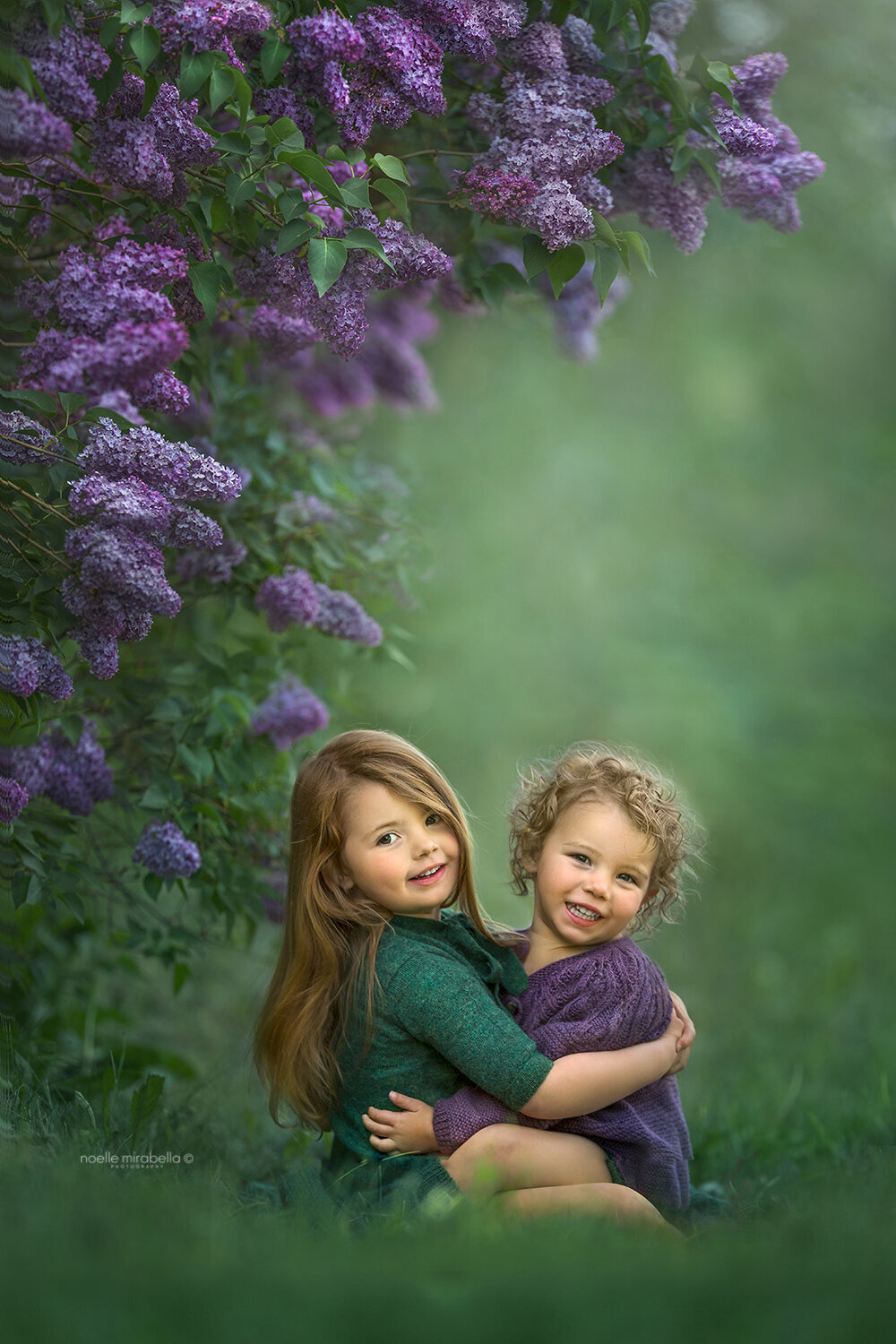 Girls in knit dresses hugging under purple lilac blossoms