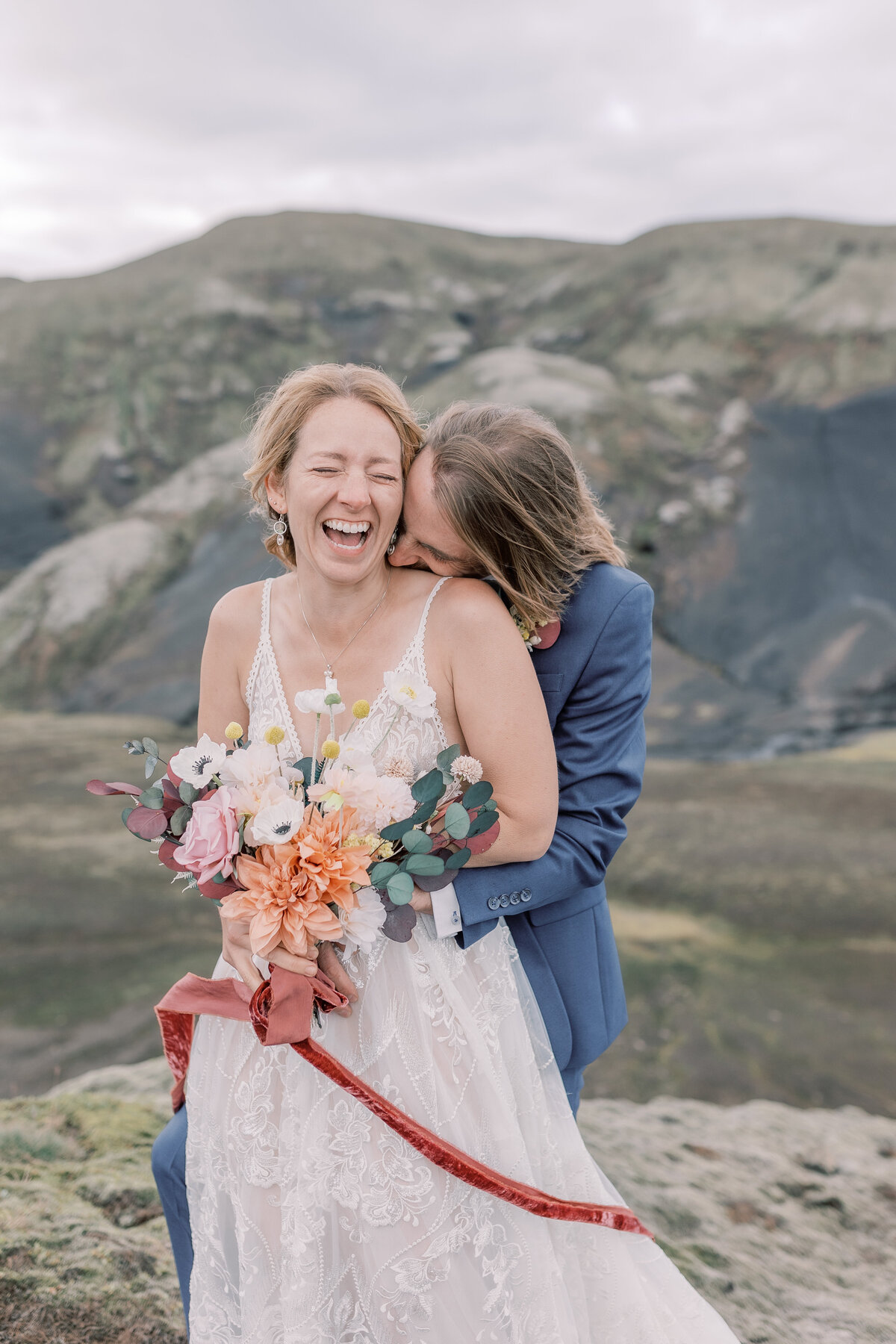 Bride holding her bouquet of dried flowers while the groom kisses her neck with the Esja mountain range in the background.