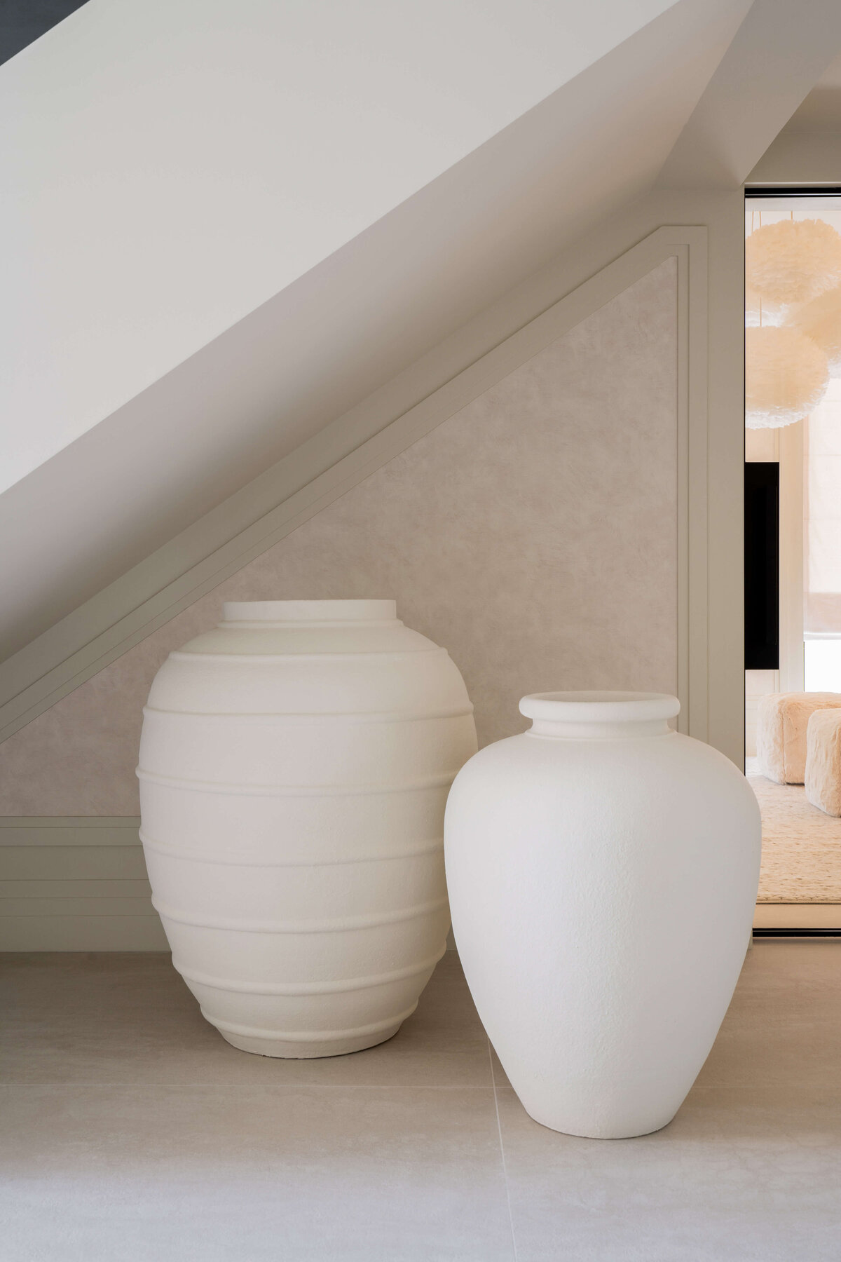 Two white vases beneath a large, modern staircase.  The vase on the left is slightly taller and has thin raised stripes. The shorter vase to the right is solid white with no texture.