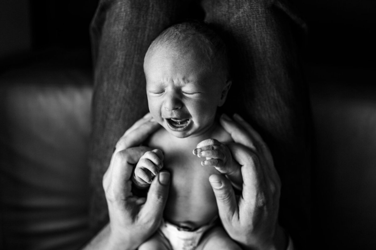 crying newborn laying in Dad's lap with his hands wrapped around baby