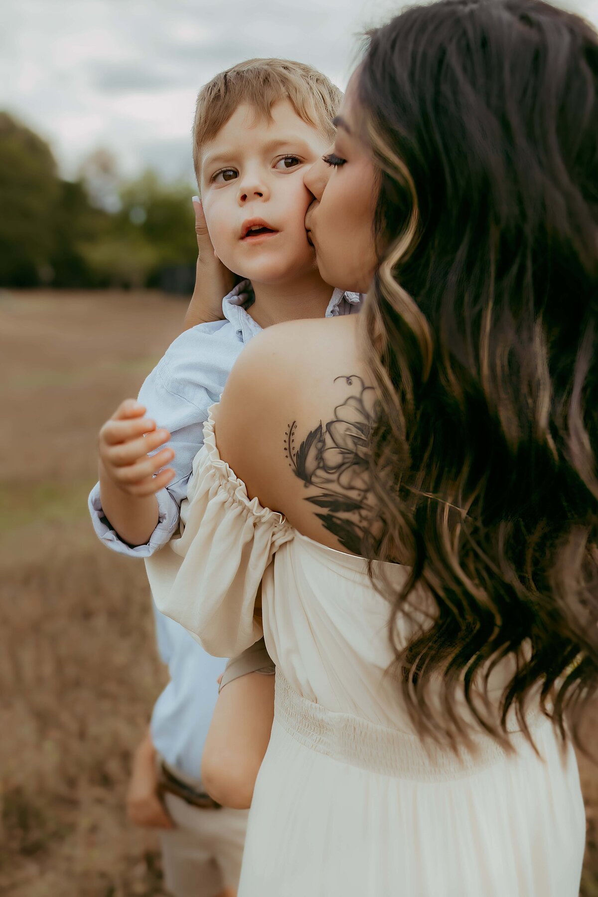 Woman kisses a young boy on the cheek while holding him. The woman has a floral tattoo on her shoulder. They are outdoors, with trees and an open field in the background.