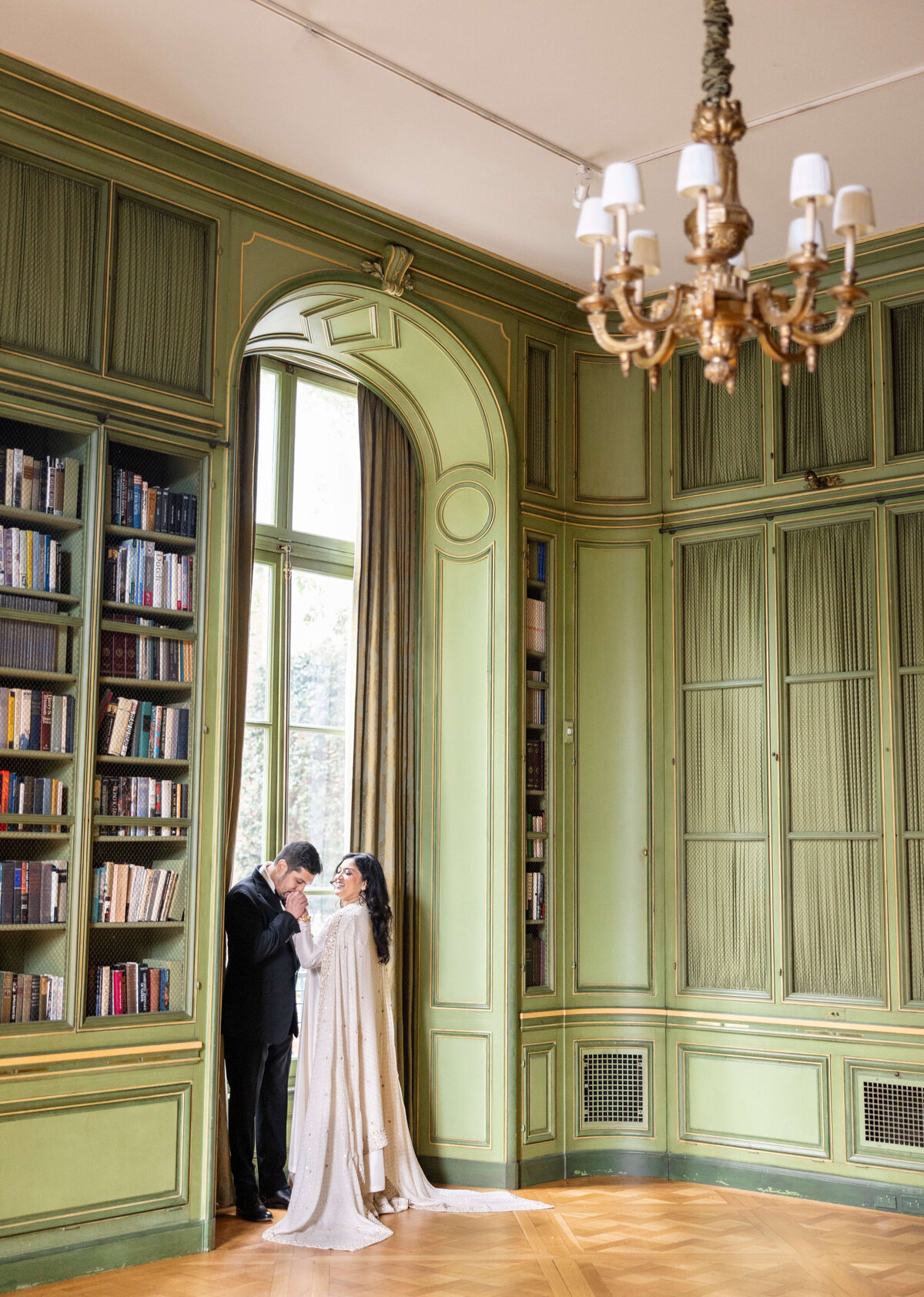 A couple stands close together, sharing an intimate moment in a grand, elegantly decorated room with tall bookshelves and a large window. The walls are painted green, and a chandelier hangs from the ceiling. Sunlight filters through the window.