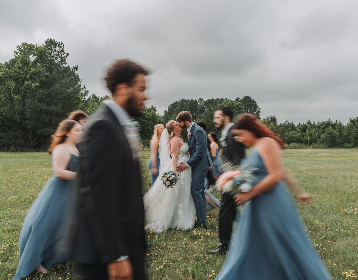A joyous wedding party, featuring the bride and groom sharing a kiss in the center, surrounded by bridesmaids in blue dresses and groomsmen, celebrating in an open field at the Hampton Roads Event Center, captured by Quinn Photography LLC.