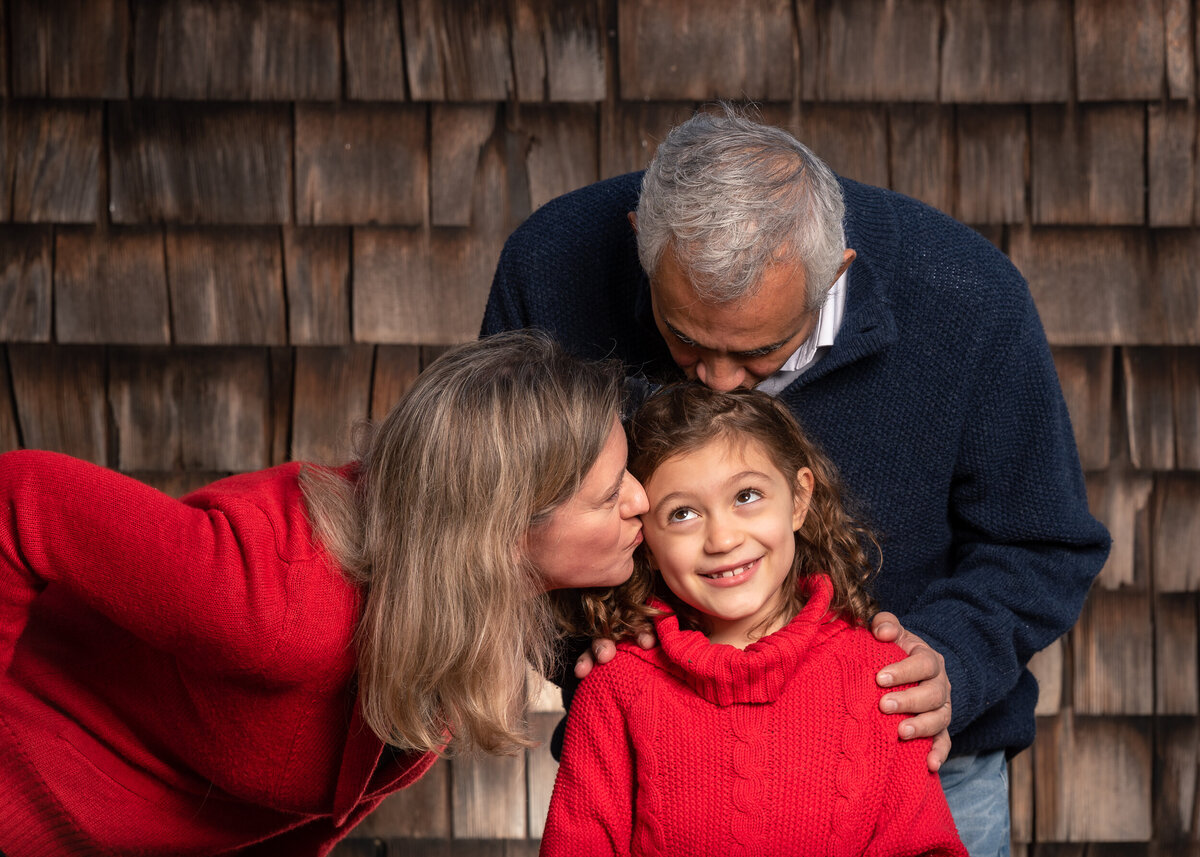 Family enjoying a photography session on a farm near Boston,  mom and dad kissing daughter in front of barn shingles