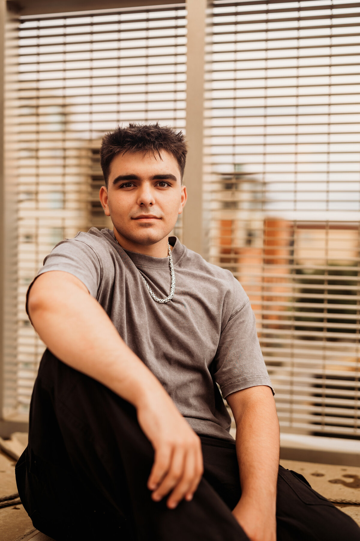High School senior boy sits on a concrete pillar posing for his senior portrait session with an urban background downtown