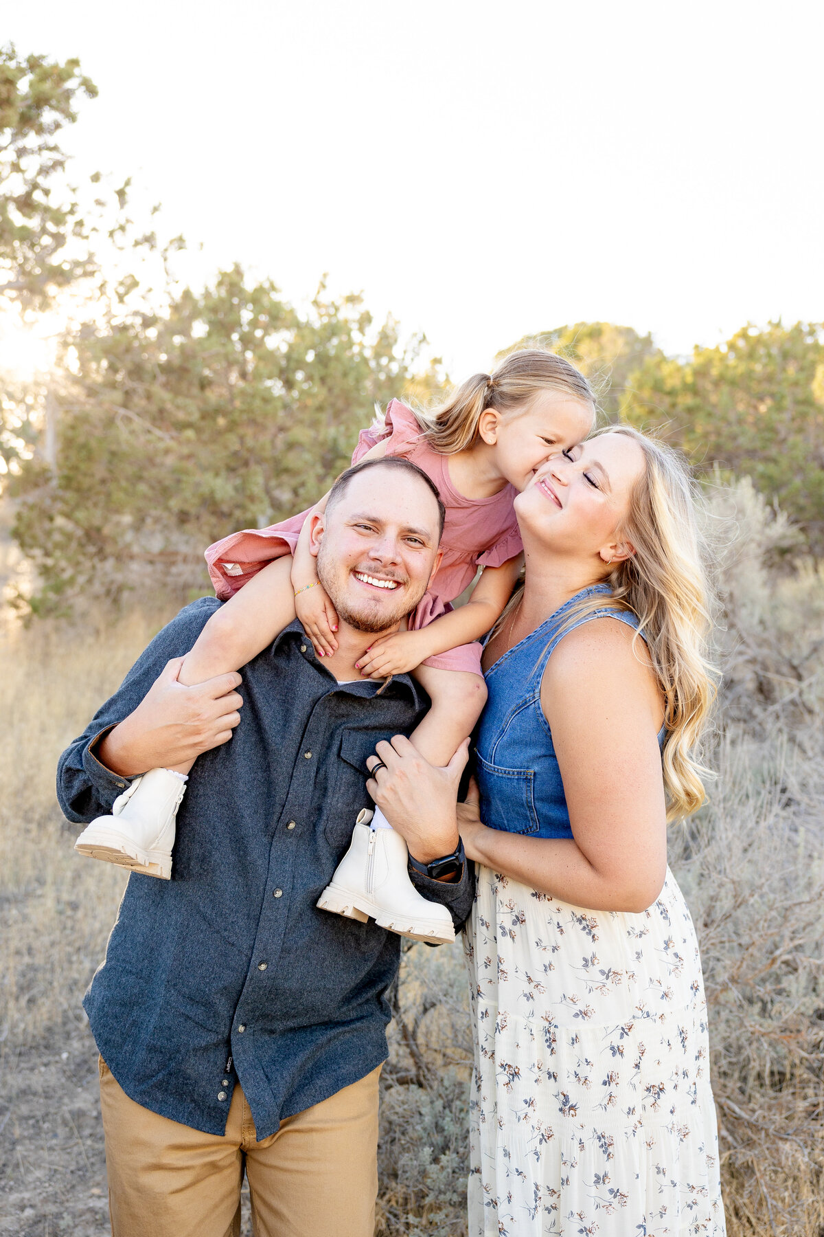 A father carries his daughter on his shoulders as she leans in to kiss her mother’s cheek. The family shares a joyful moment in the golden light of Eagle Mountain, Utah.