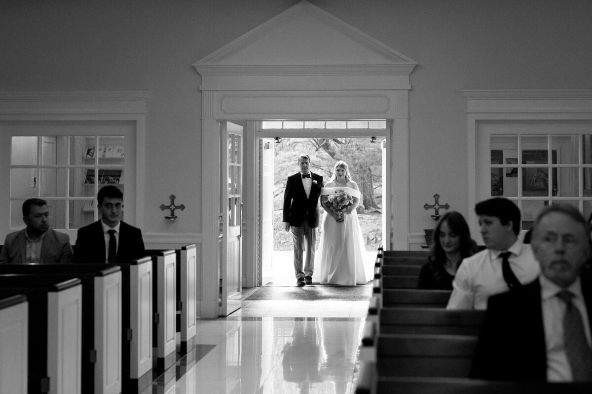 father of the bride stands in the doorway of a chapel with the bride as they begin to walk down the aisle together at the beginning of the Richmond wedding ceremony