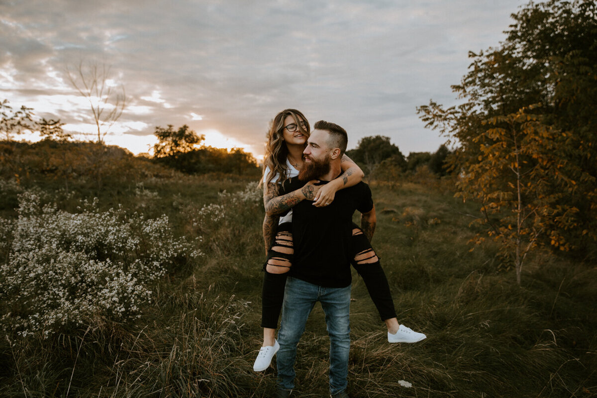 Man giving woman a piggyback through a London, Ontario field for an engagement session at sunset. Warm golden light surrounds them.
