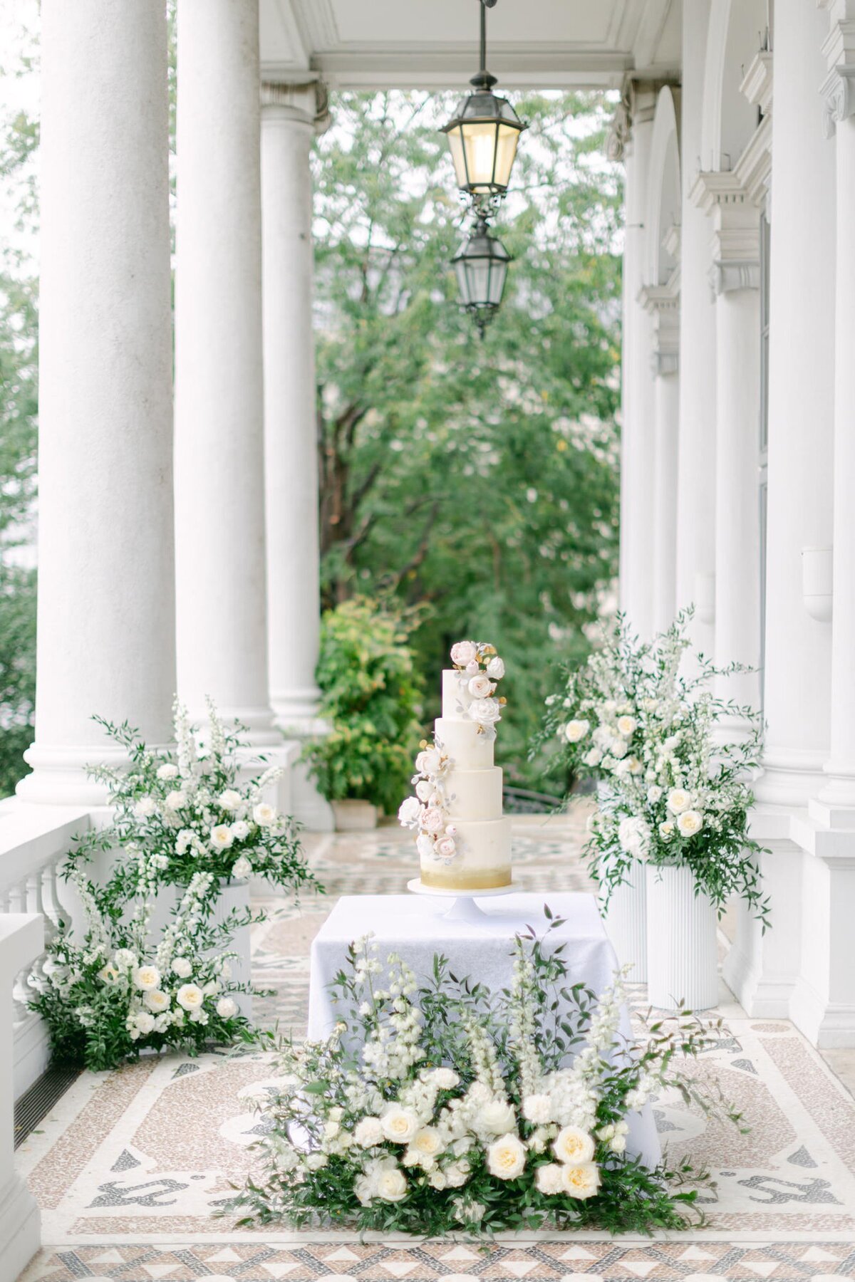 White and gold wedding cake displayed on a terrace surrounded by flower arrangements
