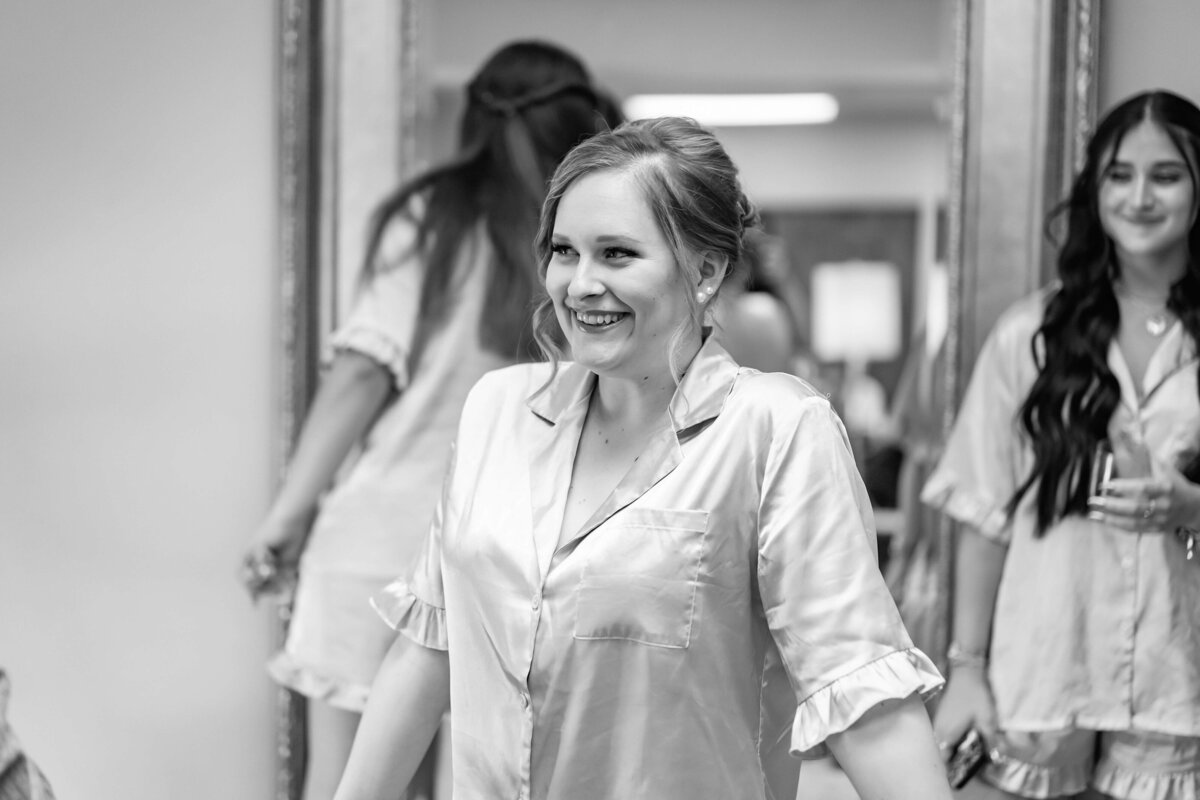 A woman in satin pajamas smiles while standing in front of a large mirror, with two other women in matching outfits in the background. The scene appears casual and joyful, likely during a pre-wedding event. The photo is in black and white.