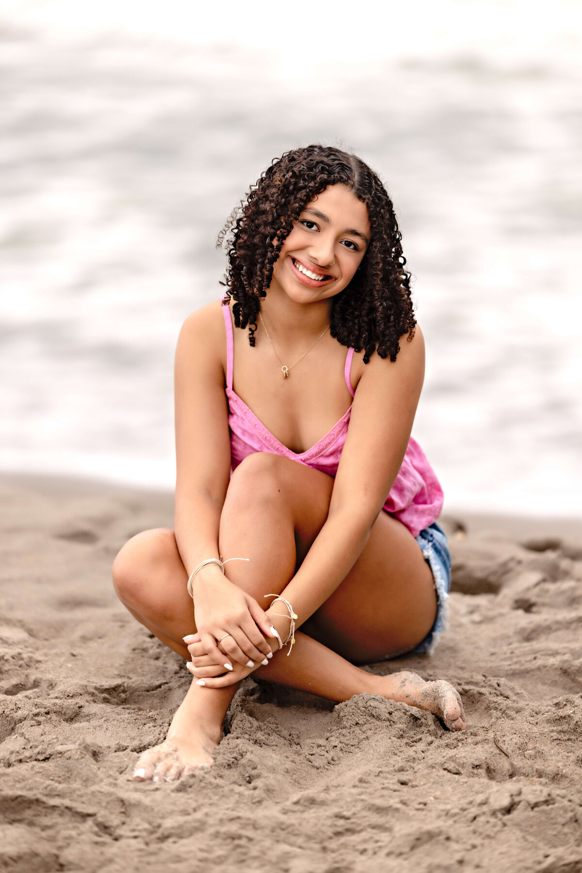 Lake Nona senior portrait of a girl sitting in the sand, legs crossed, smiling gently in soft stormy light, beach in the background.