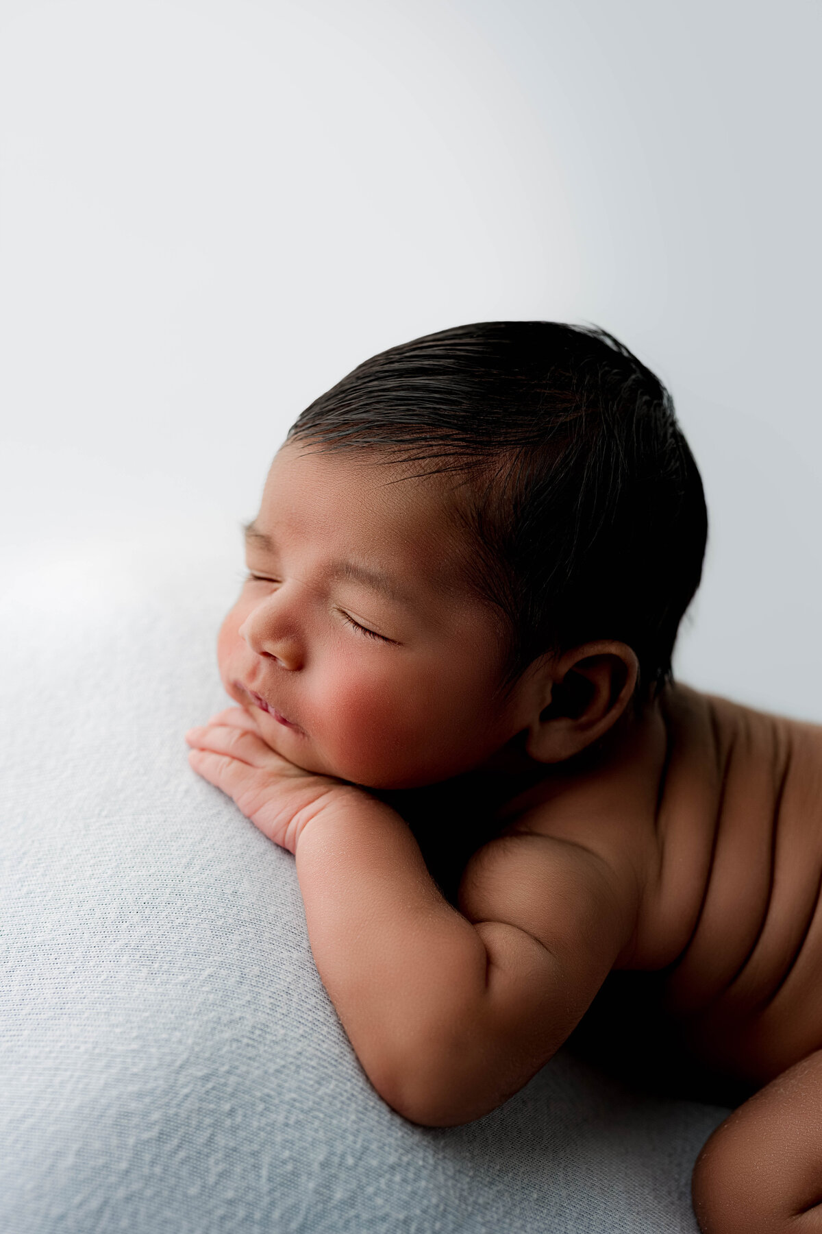 Newborn baby posed on their side with chin resting on hands, lying on soft blue fabric.