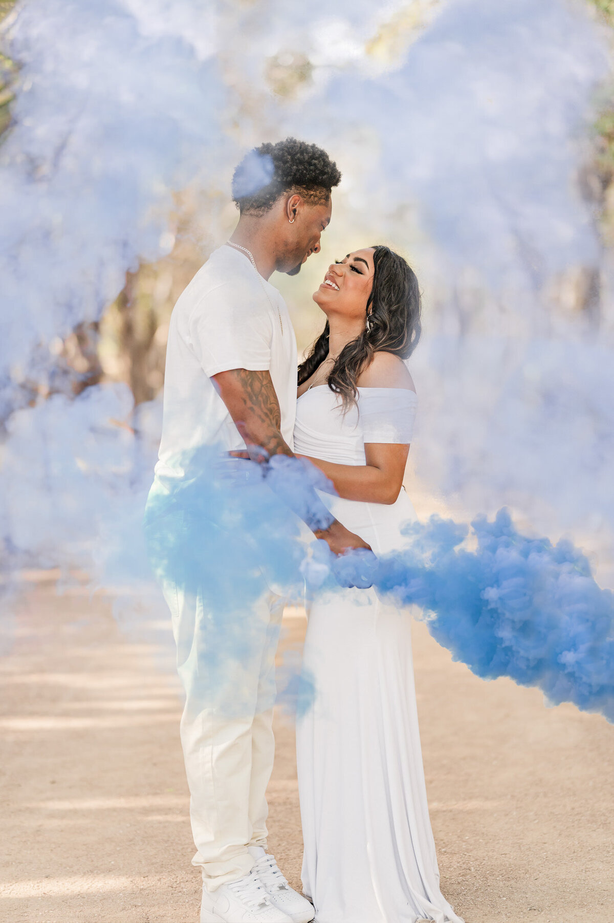Couple embraces and smiles amidst blue gender reveal smoke from maternity pictures in San Antonio.