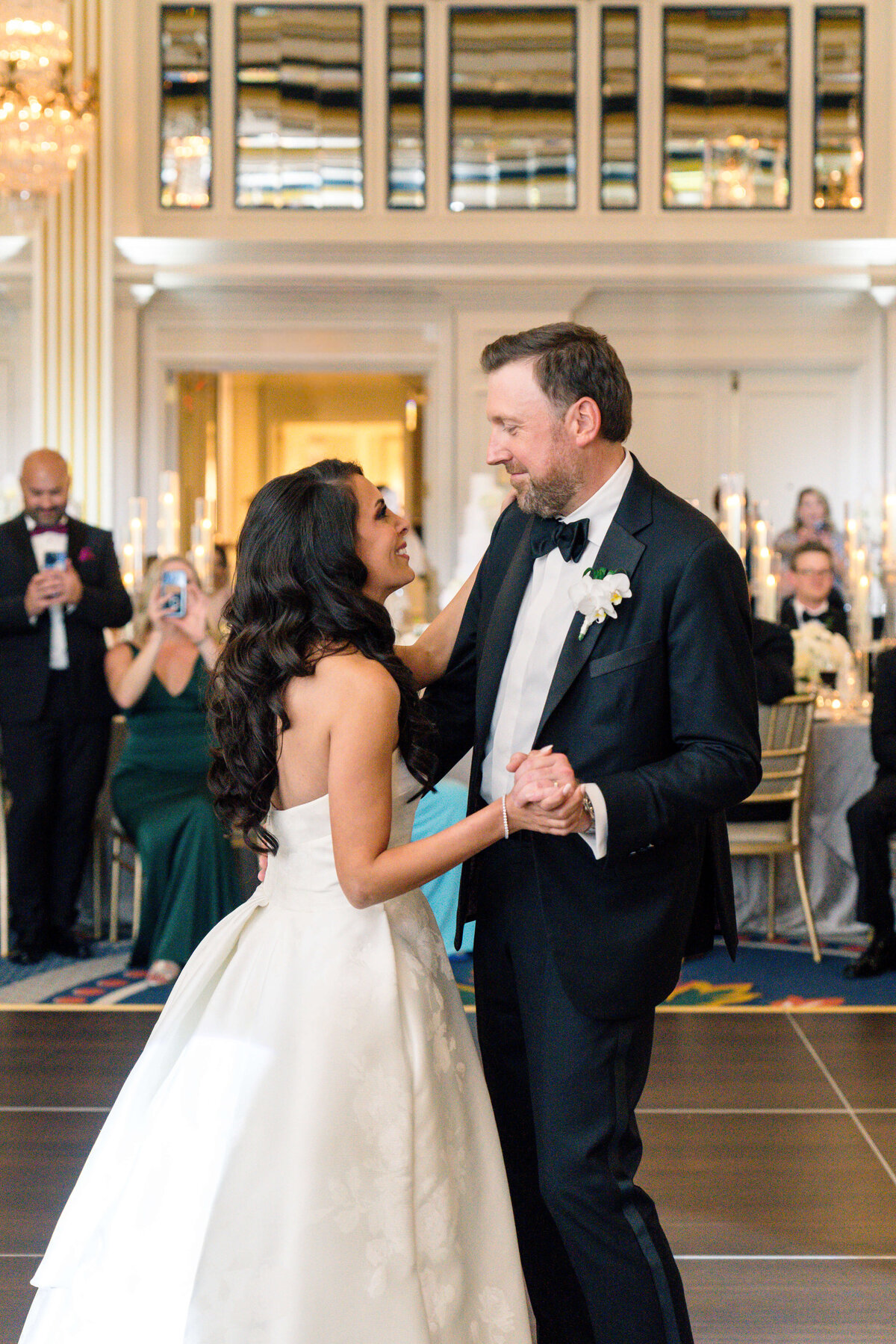 A bride and groom dance together on a ballroom floor. The bride wears a strapless white gown, and the groom is in a black tuxedo. Guests watch and smile from their seats, with elegant decor and candlelight in the background.