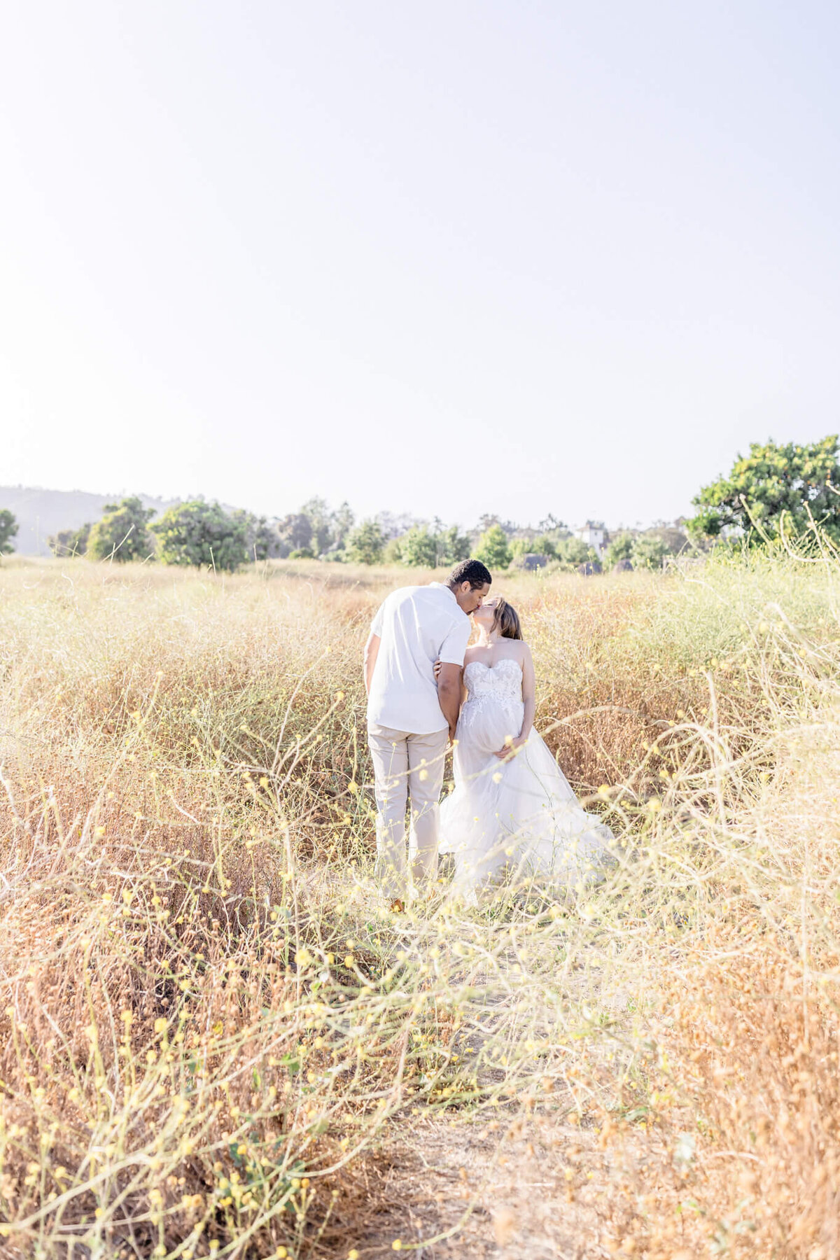 A couple in wedding attire stands closely, facing each other in an open field with tall grasses. The sky is clear, and the scene is bright and serene, with lush greenery in the distance.