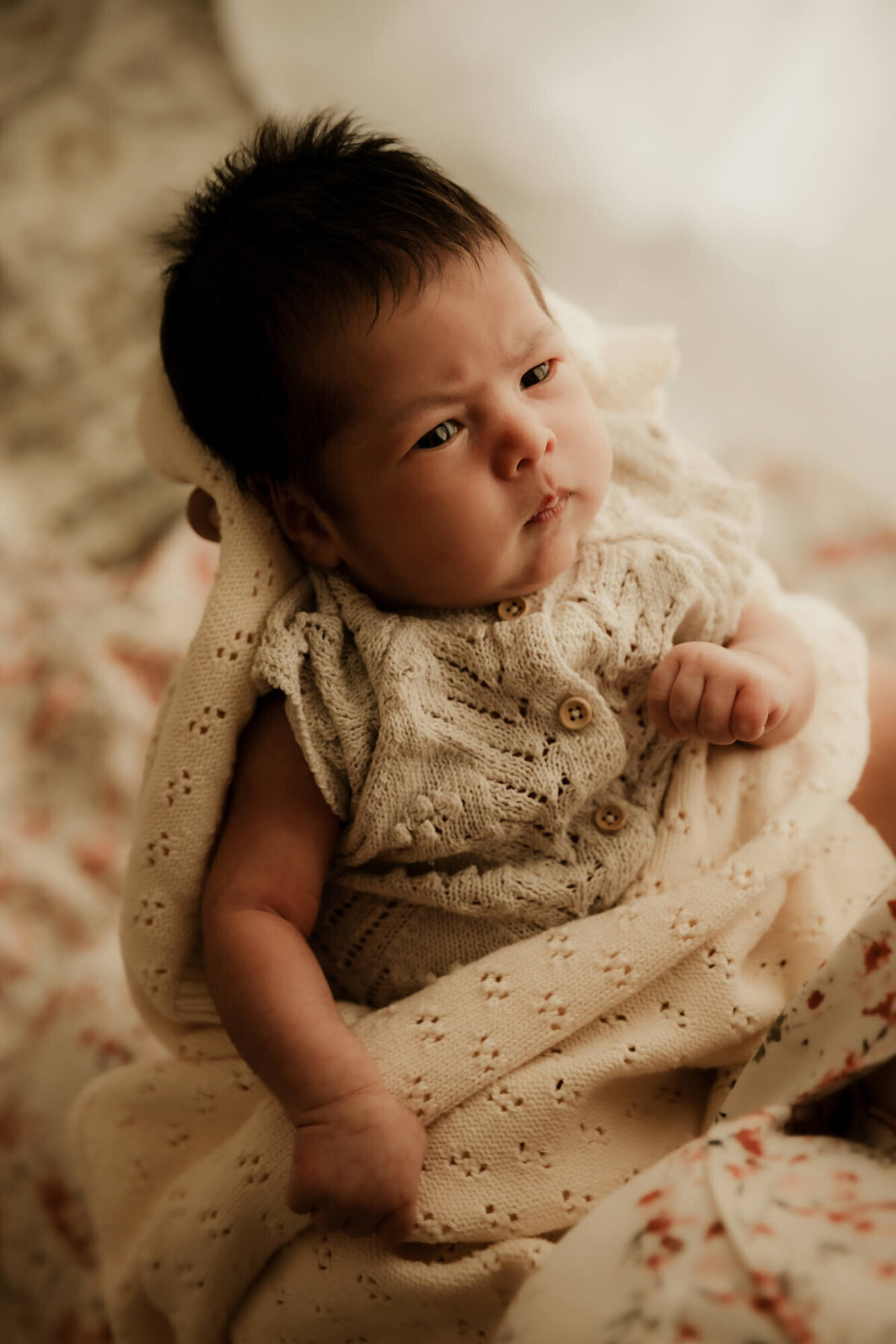 Baby with black hair wrapped in a cream colored blanket wearing a buttoned romper