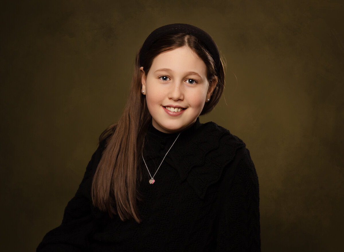 Young girl is sitting for a portrait in a brooklyn ny photo studio. She is smiling at the camera and has her hair trailing over her shoulders. Captured by Chaya Bornstein, Brooklyn Family Photographer