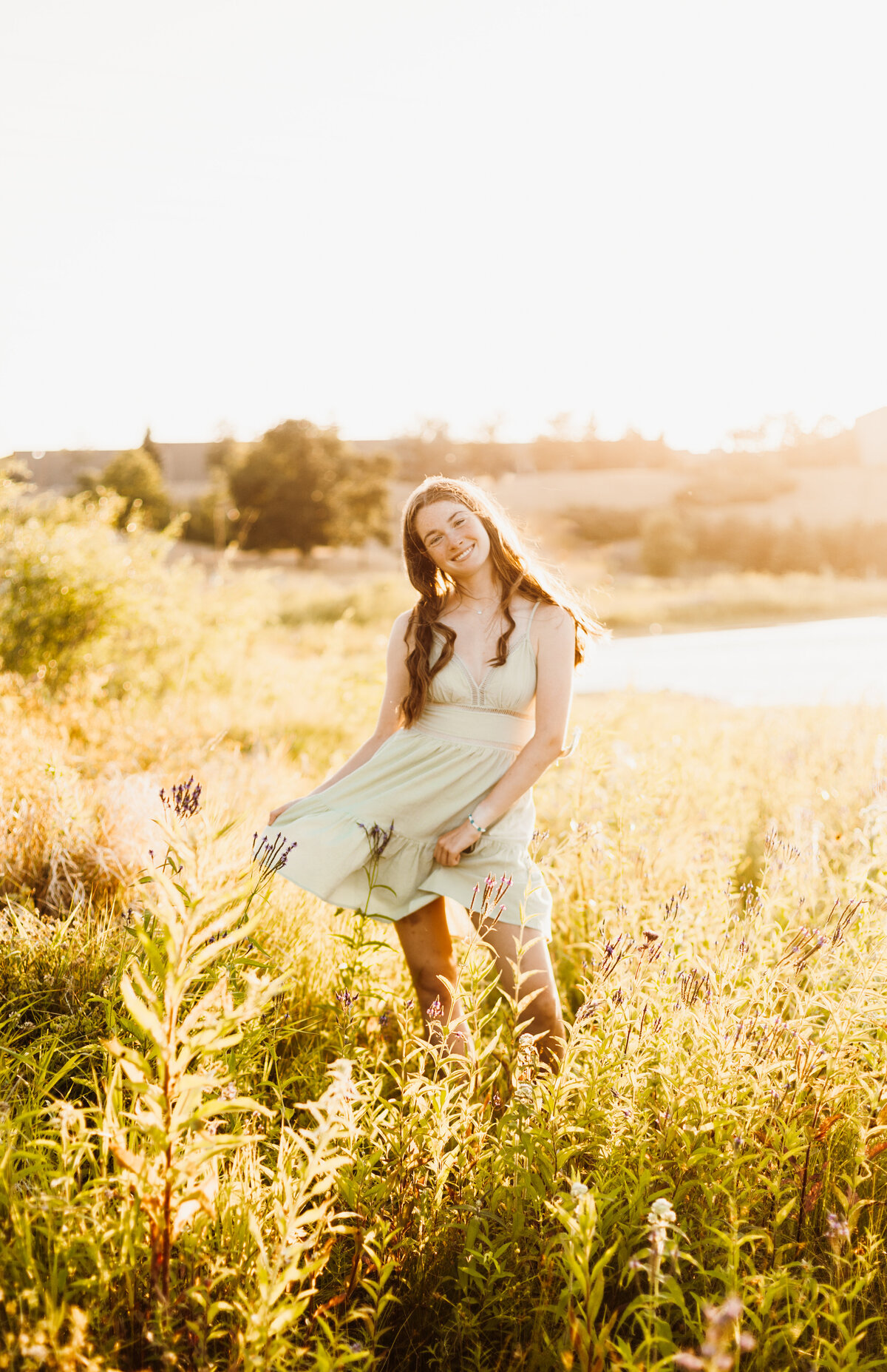 Senior girl in a light green dress posing in a grassy field with a bright sunset in the background