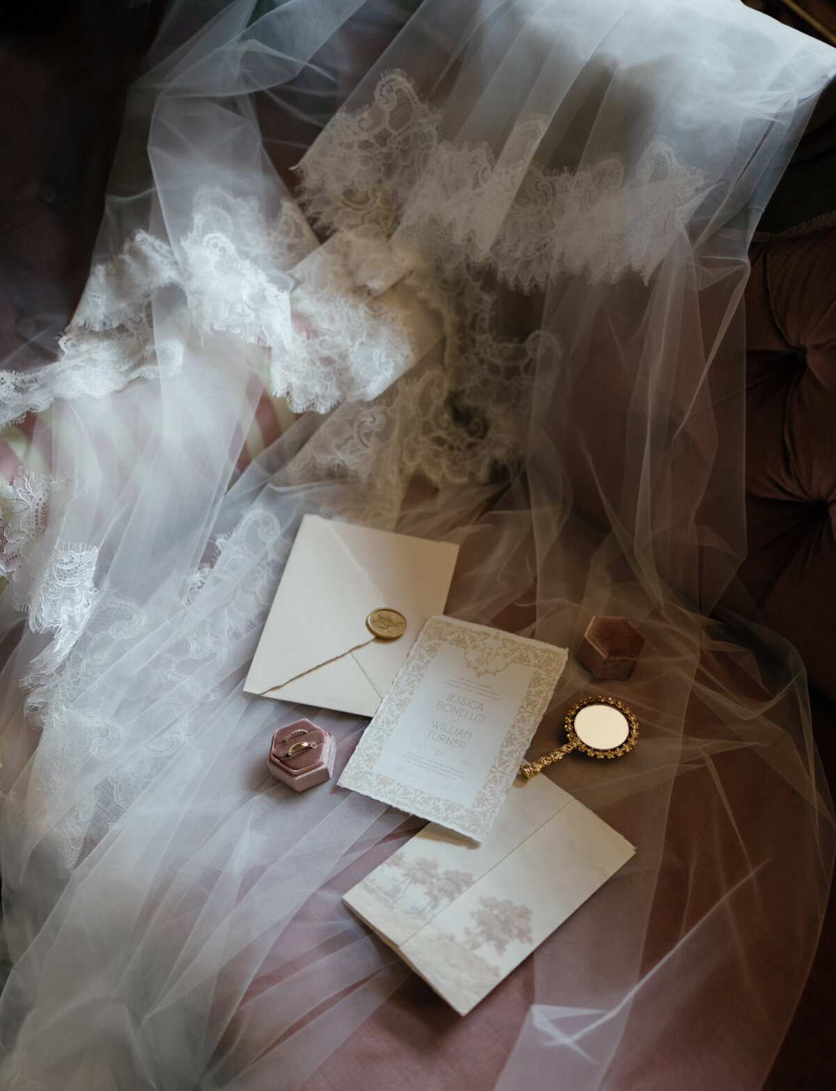soft wedding details, stationery and rings in a velvet ring box, rest in luminous light on a lace cathedral veil