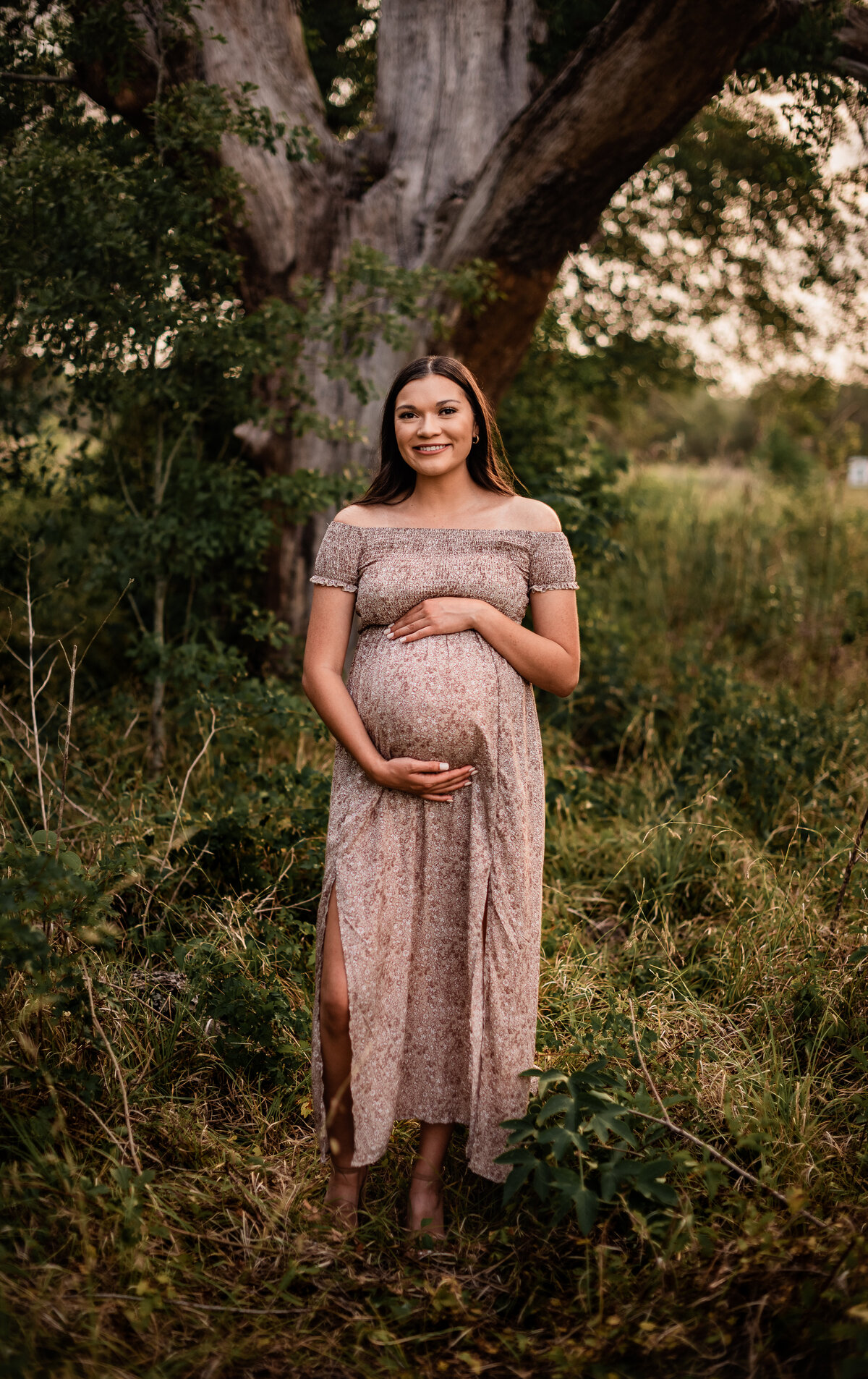 An expectant mom stands straight to the camera and cups her baby bump while she smiles toward the camera.