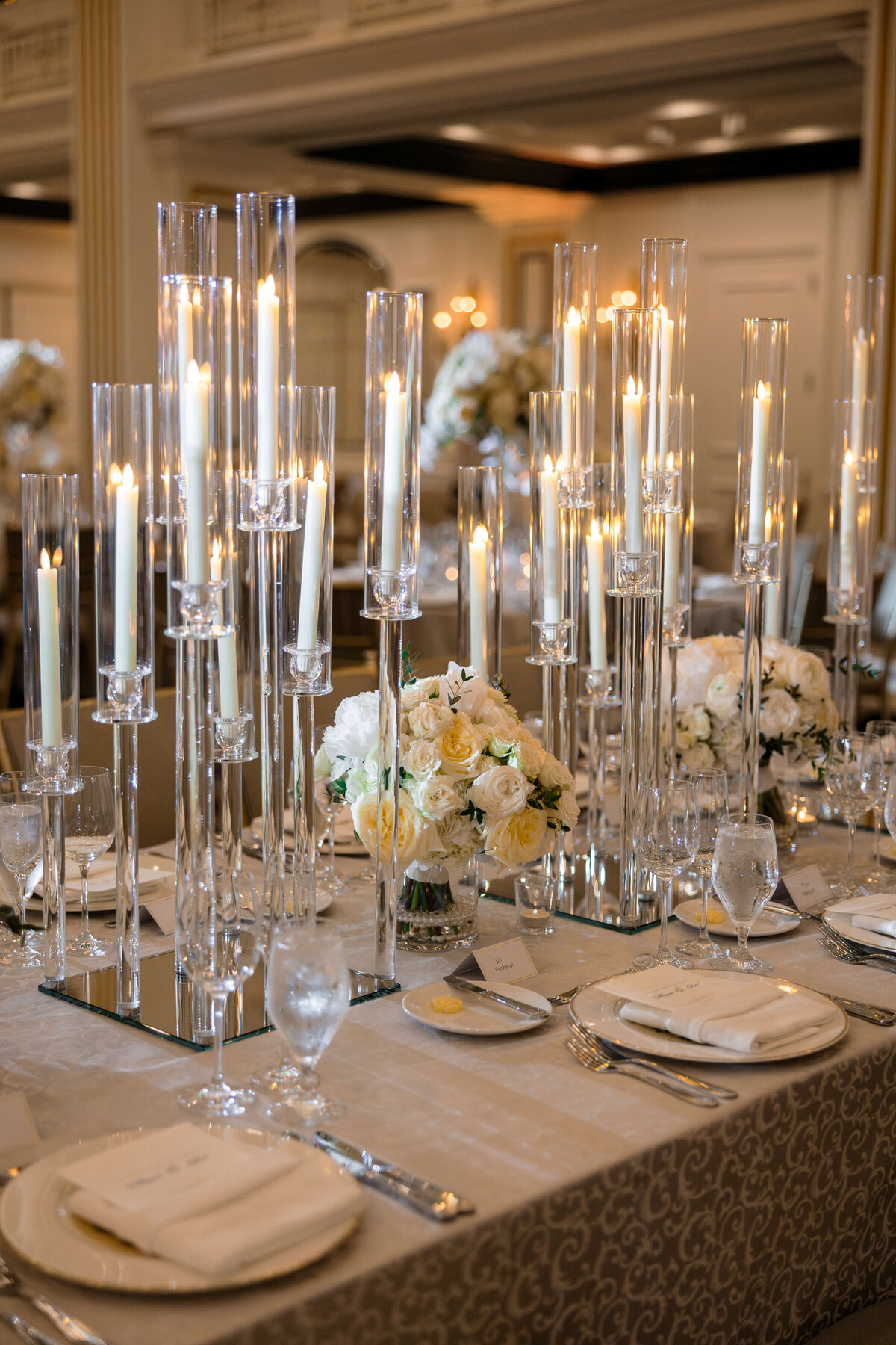 Elegant wedding table setting with tall glass candle holders, white candles, and floral arrangements of white roses. The table features neatly arranged plates, napkins, and crystal glasses, creating a sophisticated and romantic ambiance.