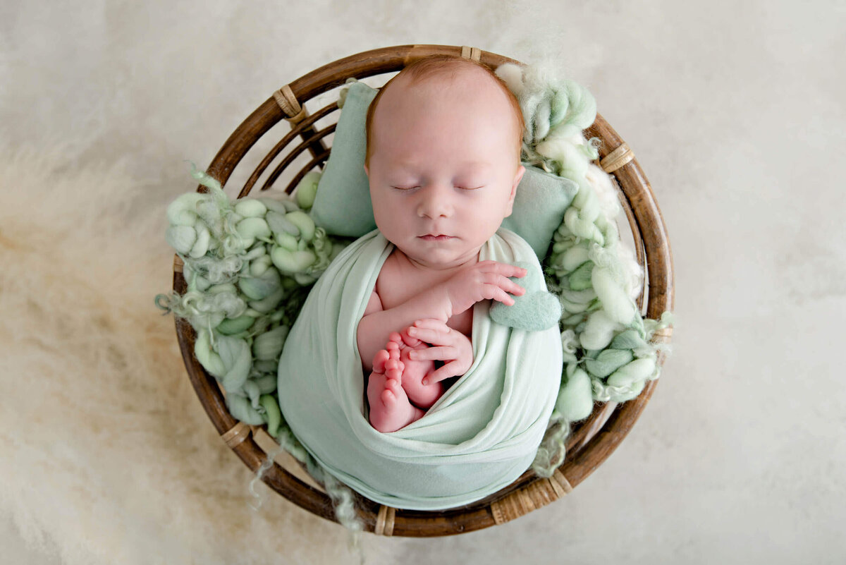 red-haired newborn boy wrapped in mint green holding a tiny green heart for his newborn photography session in Burlington.