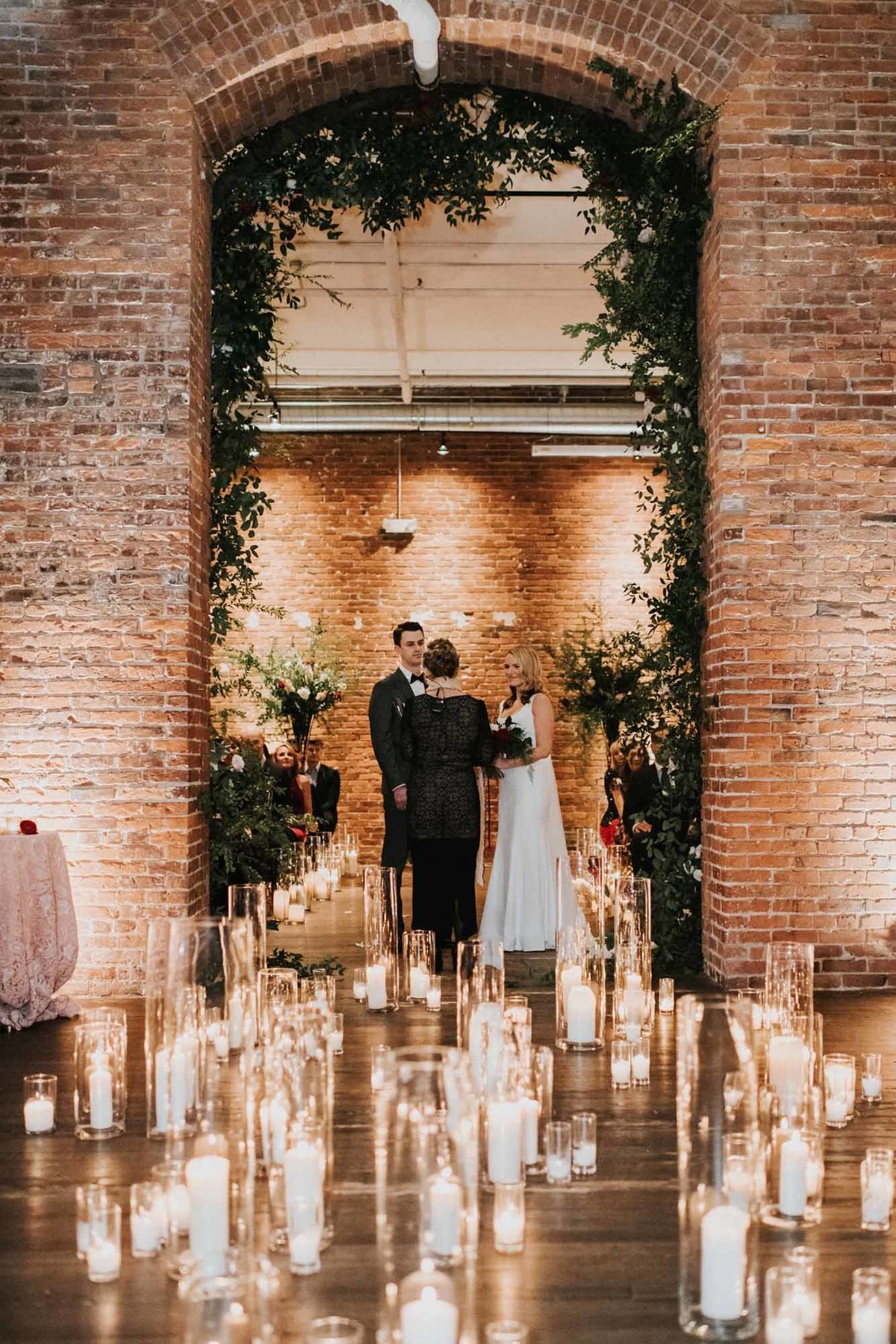 wedding ceremony under large brick arbor decorated in greenery with hundreds of hurricane candles