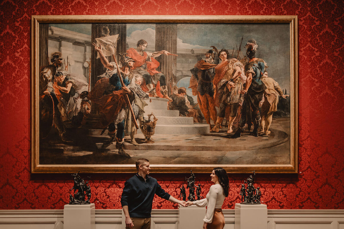 A couple holding hands in front of a large painting in a museum 