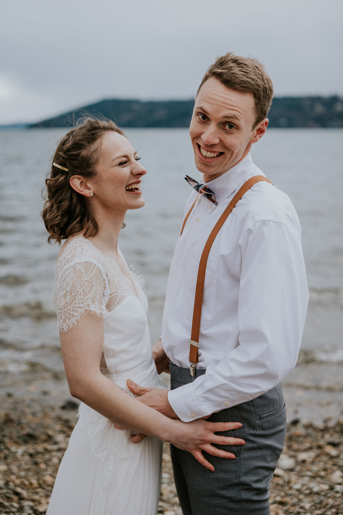 Bride and groom laugh together while standing on beach of Coeur d'Alene lake.