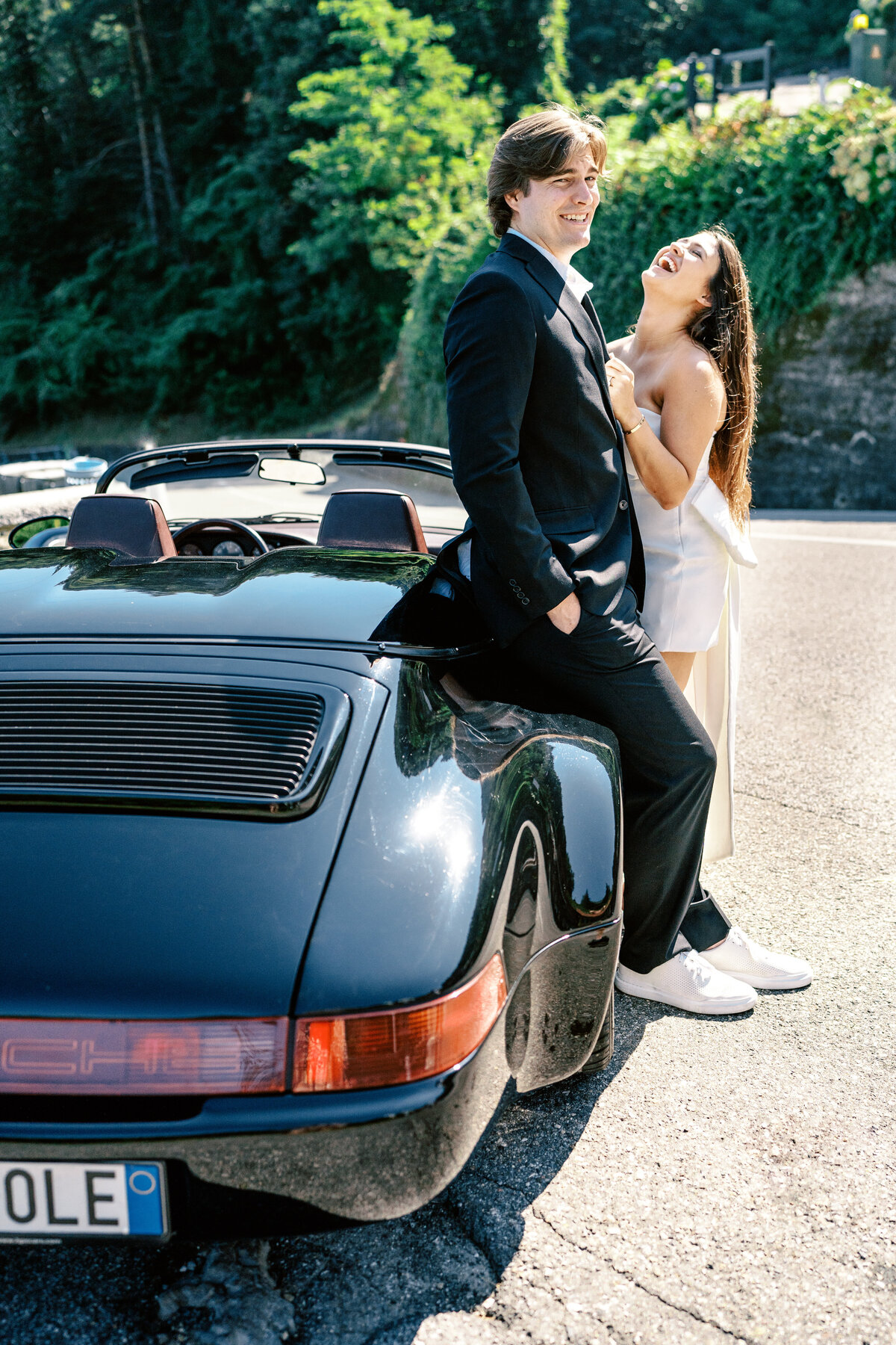 Couple laughing while standing beside black convertible Porsche for their Italy engagement session, photographed by Italy wedding photographer.