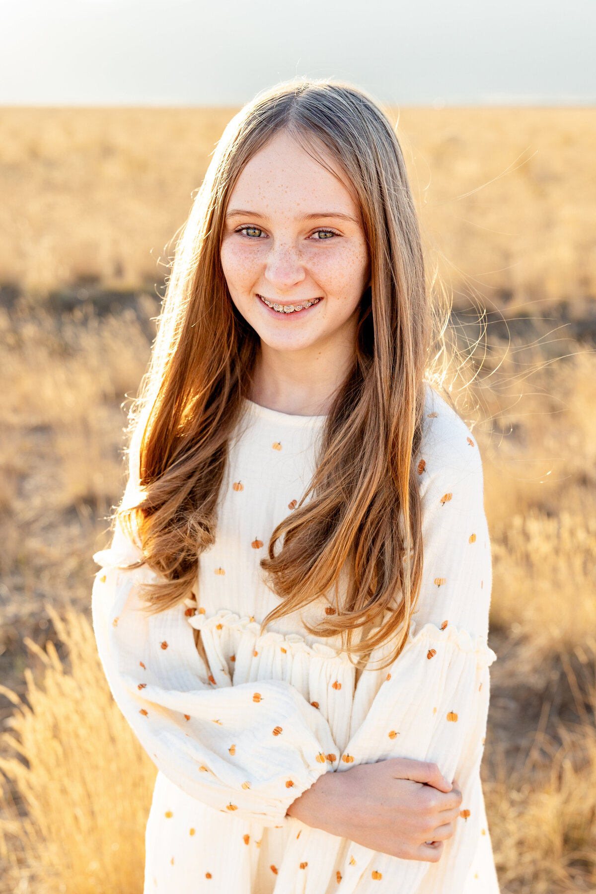 A close-up portrait of a smiling girl with long blonde hair and braces, wearing a white dress with a delicate pattern, taken in a golden field.