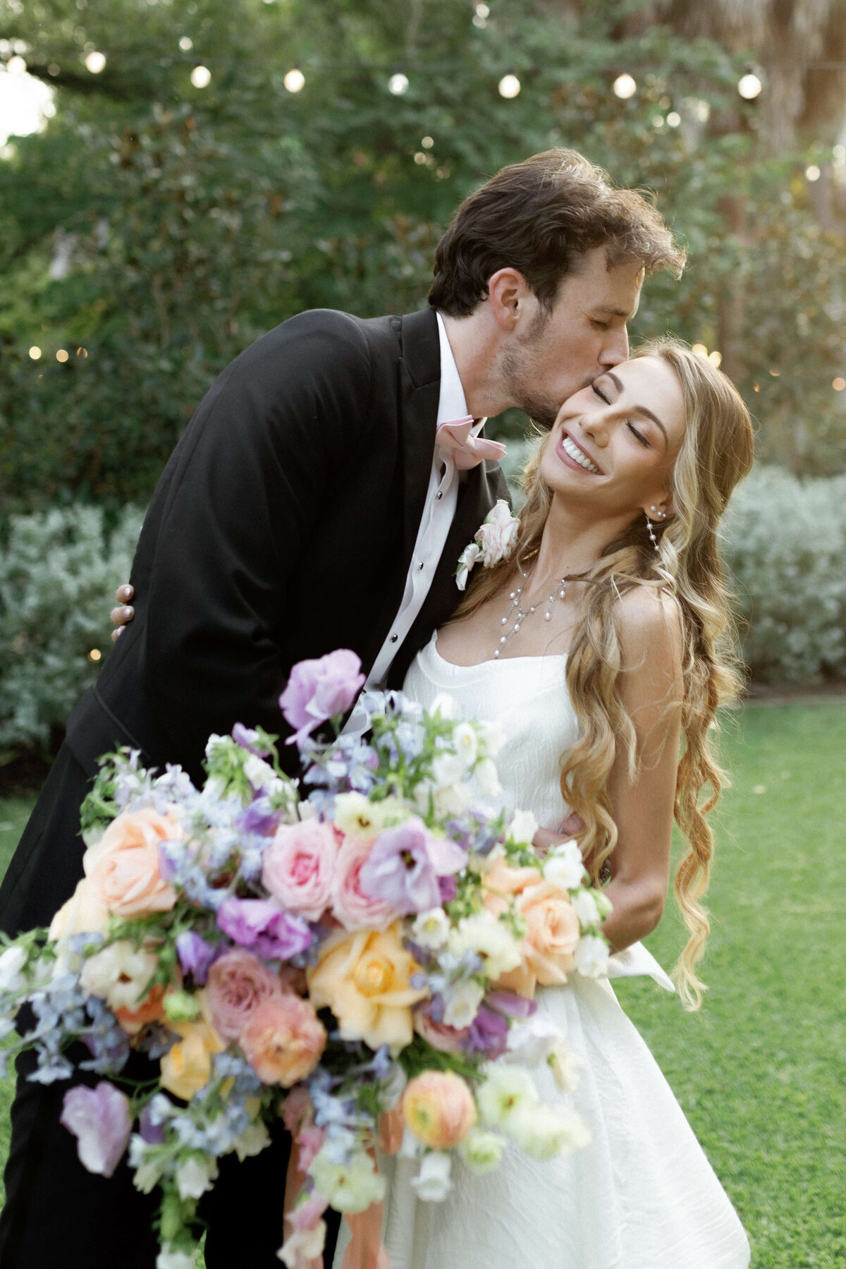 A joyful bride and groom share a sweet moment surrounded by lush greenery. The bride’s long, soft curls and radiant makeup were styled by Mistique Makeup’s wedding hair and makeup team in San Antonio. Her pastel bouquet and glowing look are perfect for spring weddings, showcasing expert bridal beauty and romantic wedding hair styling.