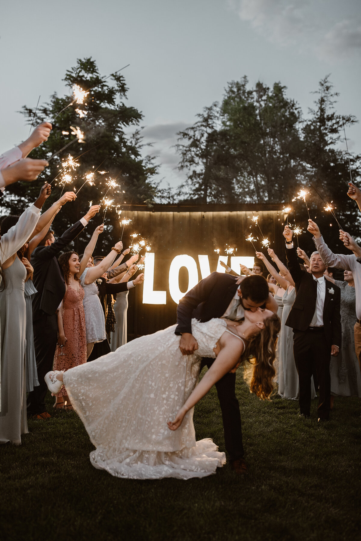 Groom dips and kisses his bride during a sparkler exit, surrounded by cheering guests and a glowing LOVE sign in the background.