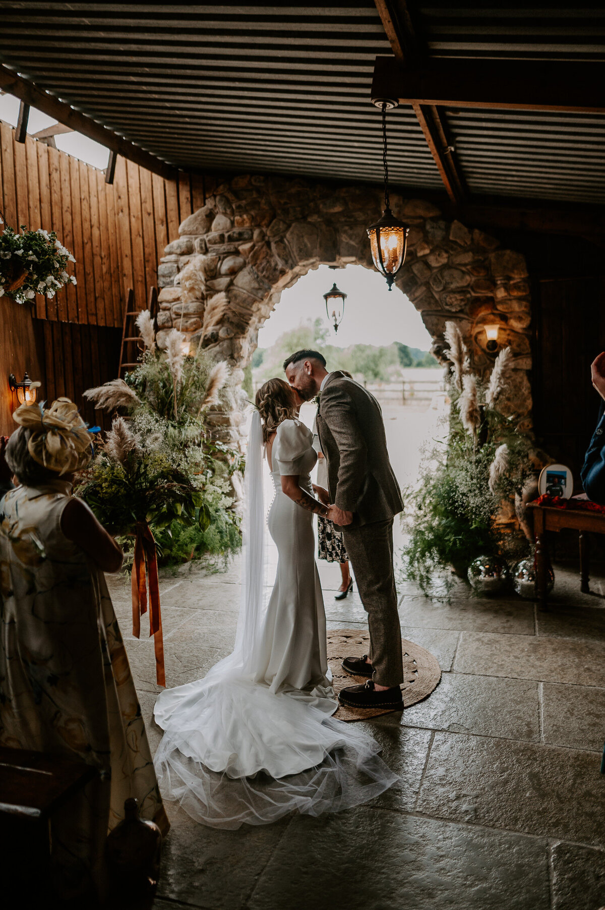 Bride and Groom have their first kiss in the stables at The Willow Marsh Farm in front of a large floral installation. The flowers are built of wild flowers and pampas grass.