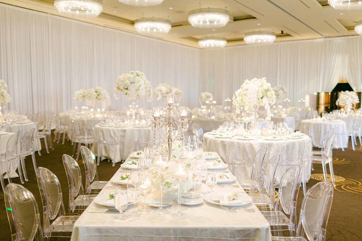 long reception table in white linen with clear ghost chairs, draped ballroom walls, tall white orchid centerpieces