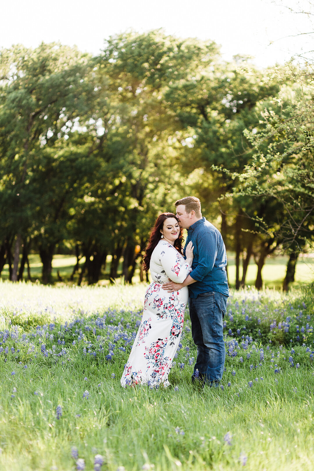 A couple holding each other close in a field of bluebonnets during their engagement session in Ennis, Texas. The woman on the right is receiving a kiss on her temple and is wearing a long white dress covered in intricate, colorful flowers. The man on the right is wearing a blue checkered dress shit and jeans.