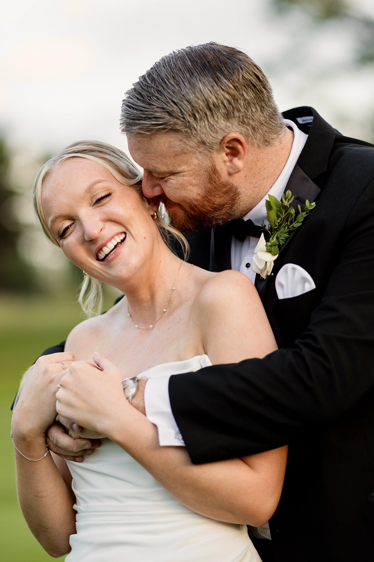 A groom embraces and kisses the cheek of a smiling bride outdoors on their wedding day. Both are dressed formally, with the groom in a black tuxedo and the bride in a white strapless dress.
