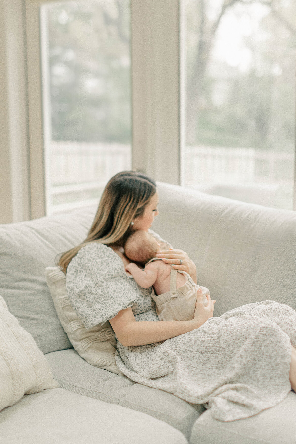 Mother sitting on a couch by a large window, cradling her newborn baby boy during an in-home lifestyle session in Kansas City.