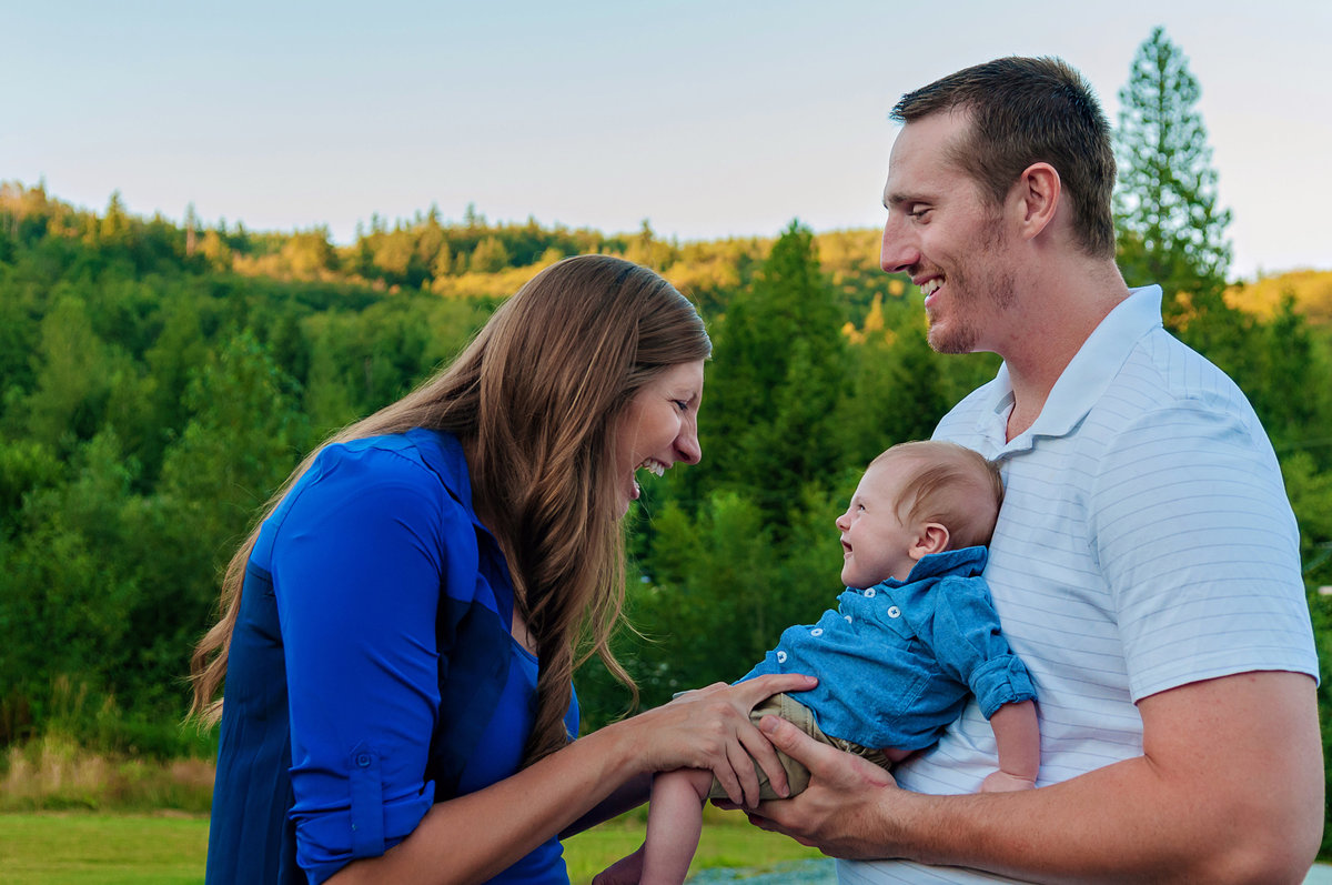 Dad holding baby and mom laughing with the baby, all dressed for summer outdoors with beautiful green trees in the background