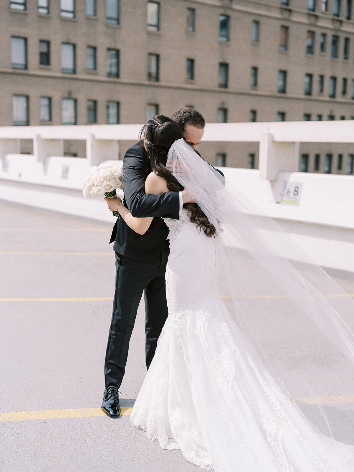 A bride and groom embrace each other outdoors in an urban setting, capturing the essence of a classic Calgary wedding. The bride, adorned in a long white dress and veil, cradles her bouquet of white flowers as they share this timeless moment.