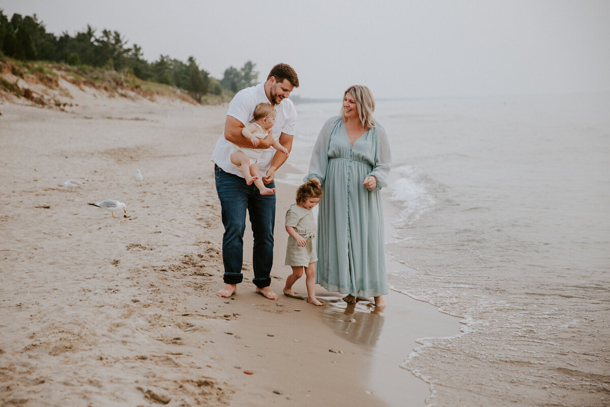 Family photoshoot at Grand Bend beach with top photographer. The family is standing in the shoreline. Mom is holding the toddler's hand, who is dipping her toes in the water. The Dad is holding the infant. Mom and Dad are smiling at each other for a candid photo.