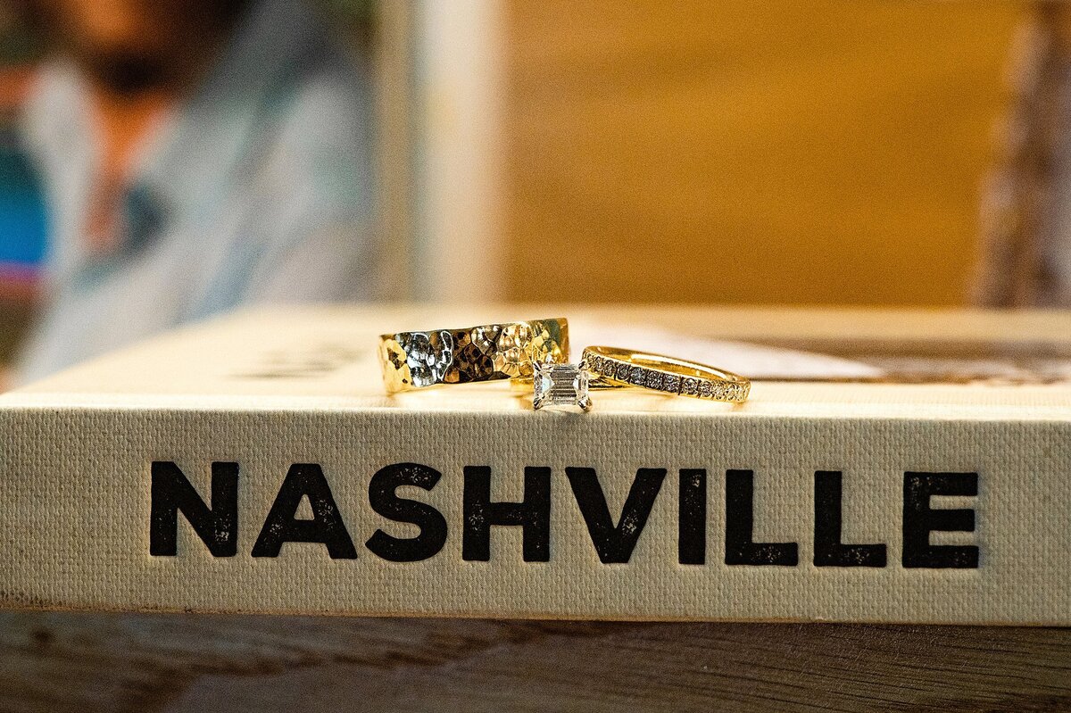 A hand hammered men's wedding band sits beside a woman's diamond eternity wedding band. Betweek both wedding rings is a square cut diamond on a gold band. The rings are sitting on top of a coffee table book titled NASHVILLE