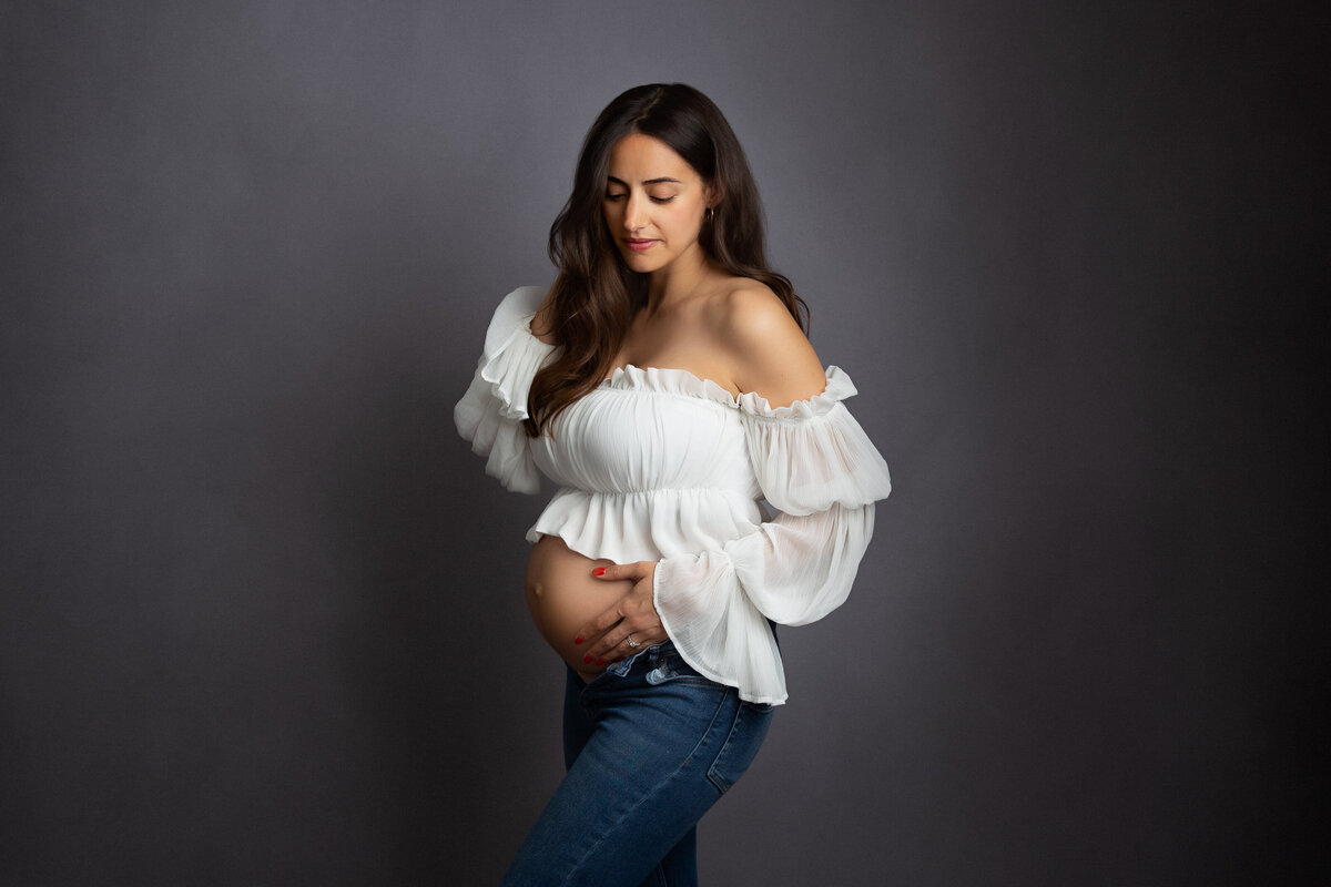 Pregnant woman wearing  blue jeans and a white chiffon crop top looking tenderly at her belly..