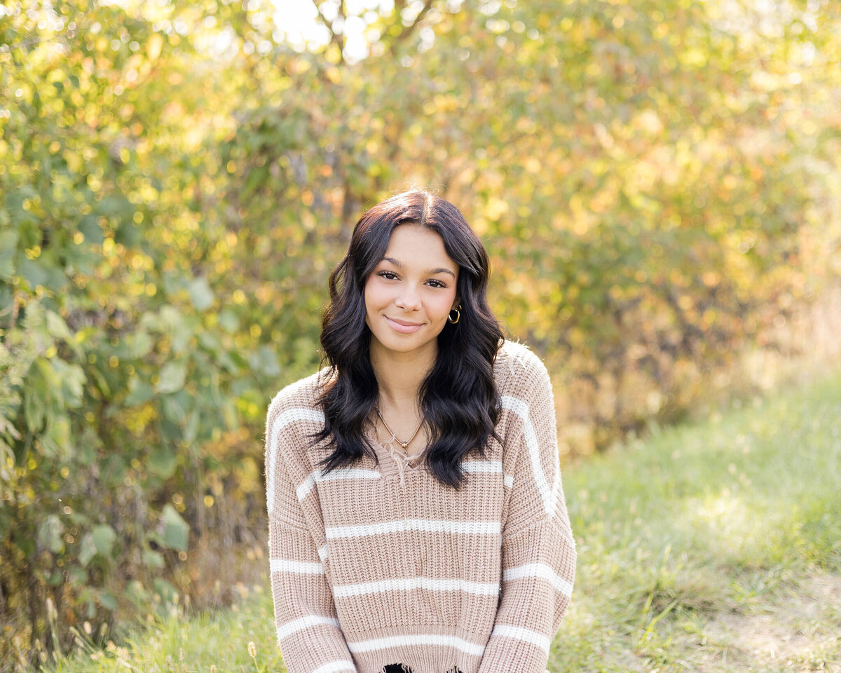 high school female sitting in grass with trees behind her, smiling at the camera, wearing black leggings, wearing tan and white striped long sleeve shirt